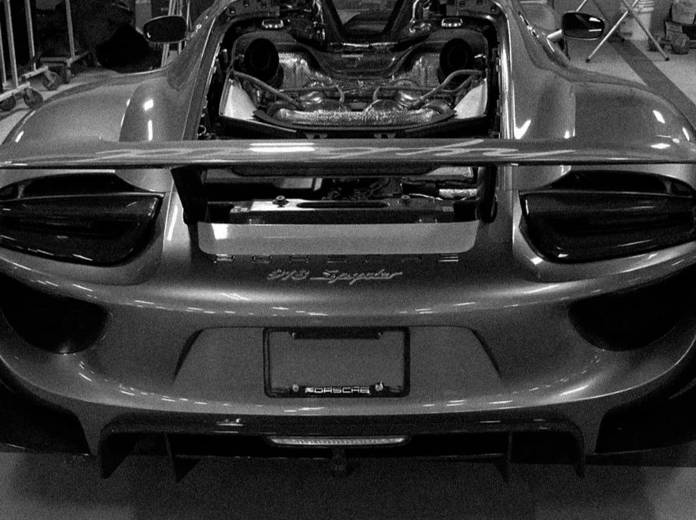 Black and white photo of the rear of a Porsche 918 Spyder sports car with the engine removed, showing the engine bay, exhaust system, and rear spoiler.
