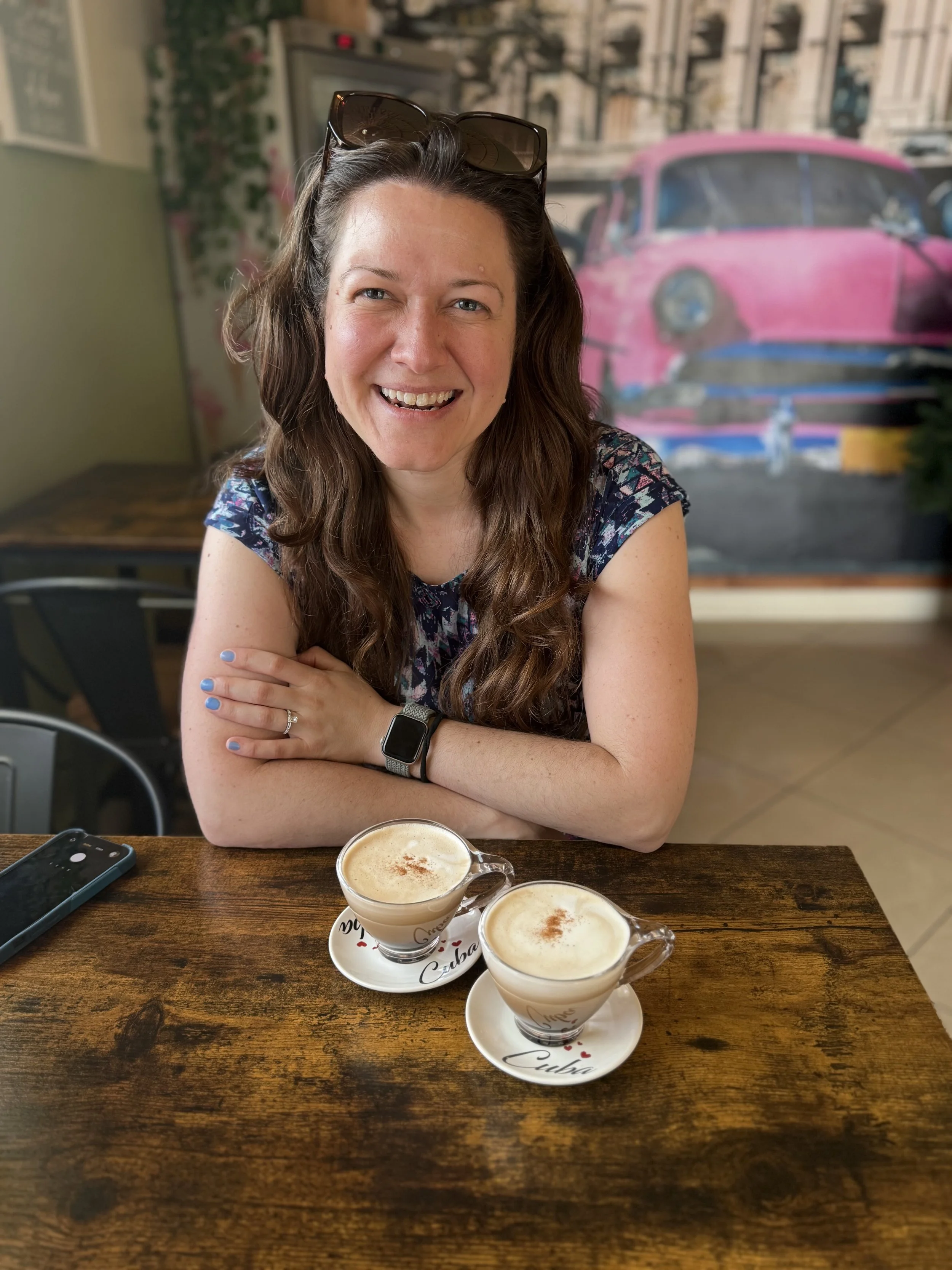 A woman with long brown hair wearing a floral shirt and sunglasses on her head, smiling and sitting at a wooden table with two cups of mocha with cinnamon on top. She is crossing her arms, and on her left wrist, she is wearing a smartwatch. There are a smartphone and a napkin with the word 'Cuba' written on it in front of her. The background shows a painting of a pink vintage truck and some garden furniture inside a cozy cafe.