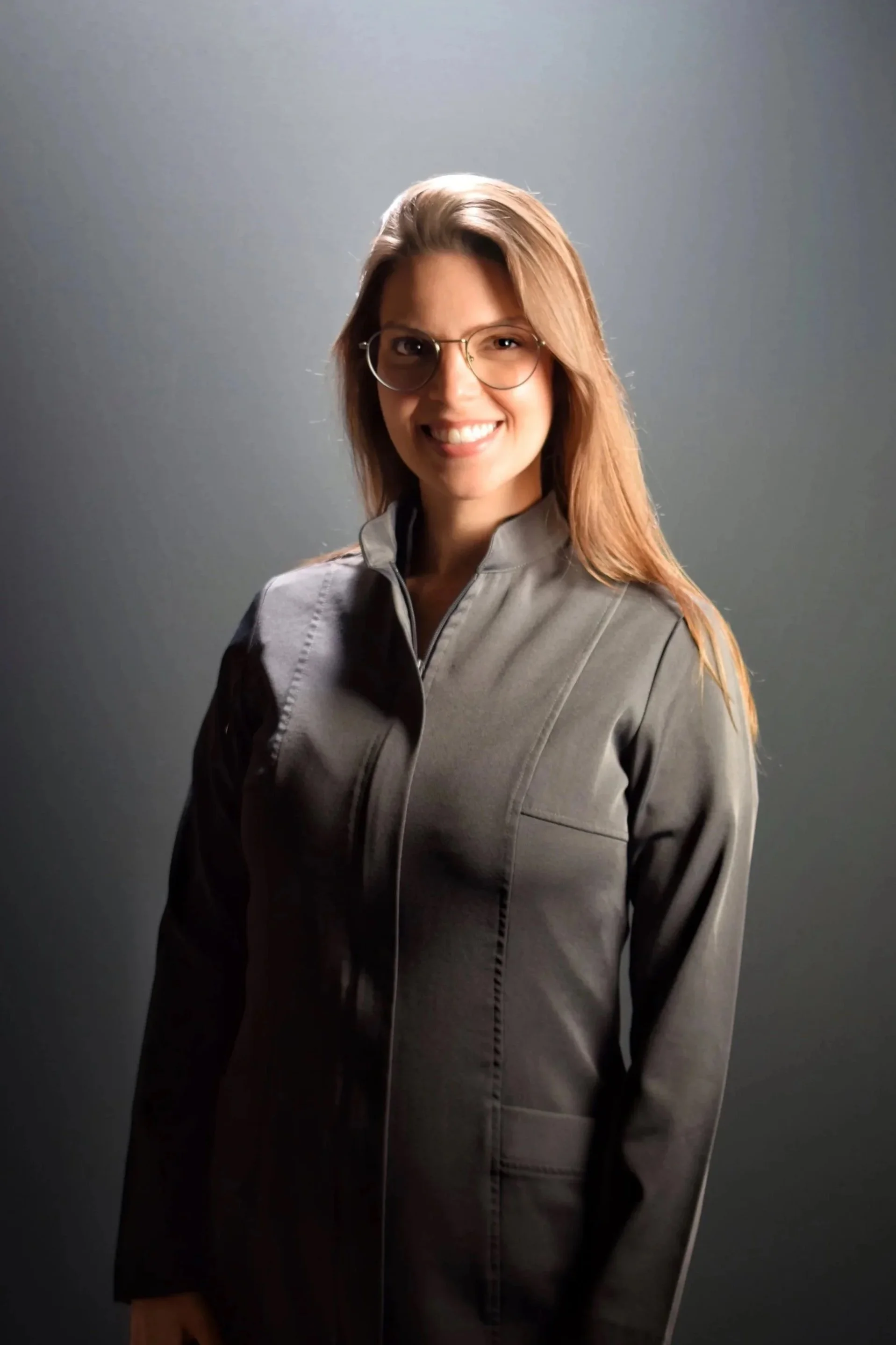 Mulher sorridente usando óculos e jaqueta cinza escura, posando contra fundo cinza.