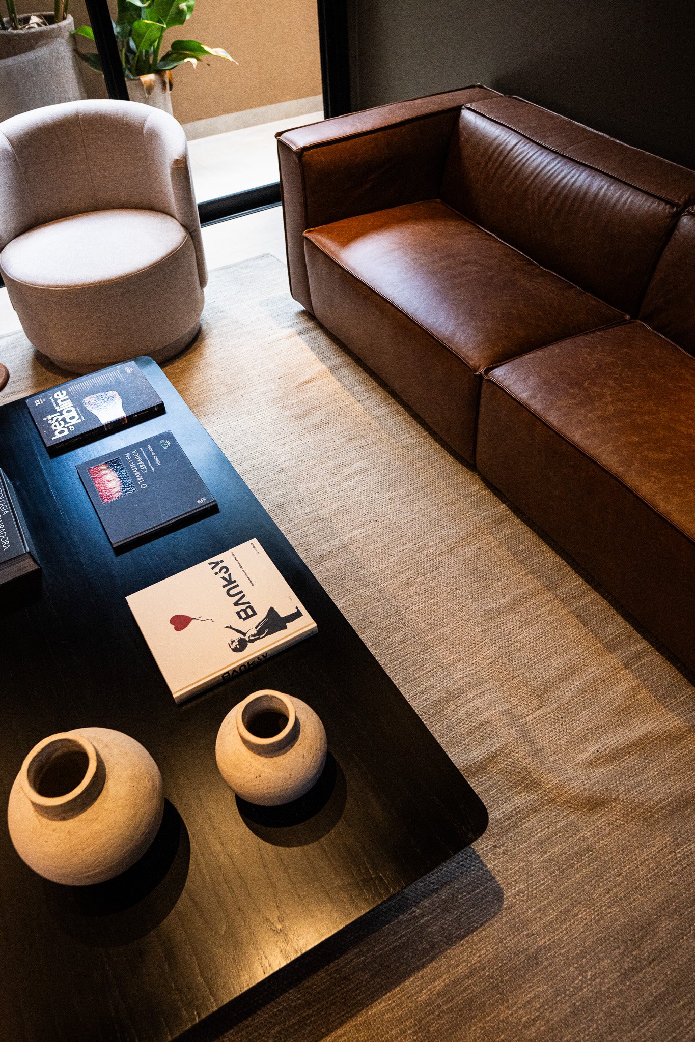 Sala de estar com sofá de couro marrom, cadeira de canto cinza, mesa de centro preta com livros e vasos de cerâmica, tapete bege, e uma porta de vidro ao fundo com plantas ao lado.