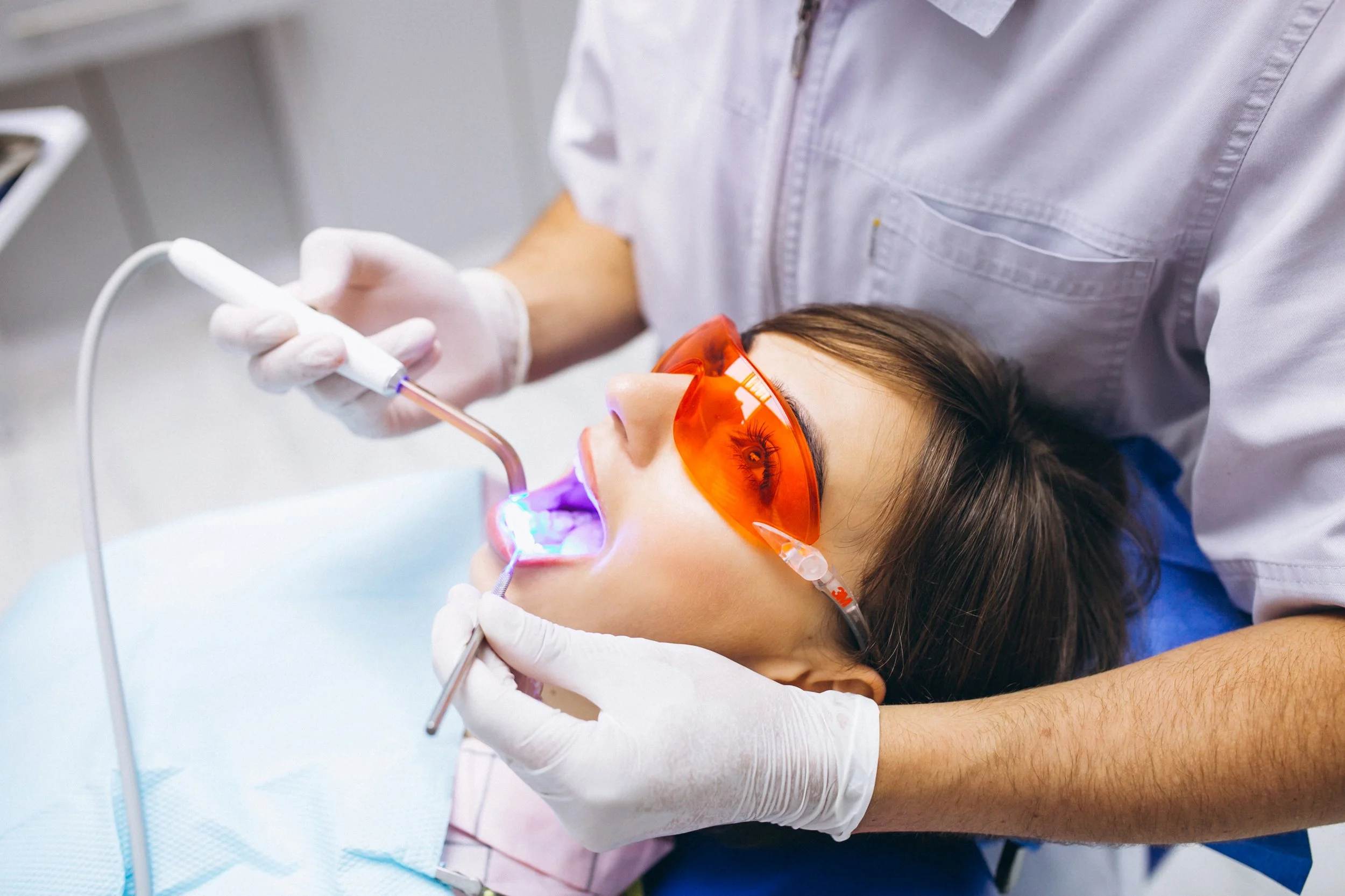 Dentista realizando procedimento odontológico em paciente, que usa óculos de proteção laranja, em consultório.