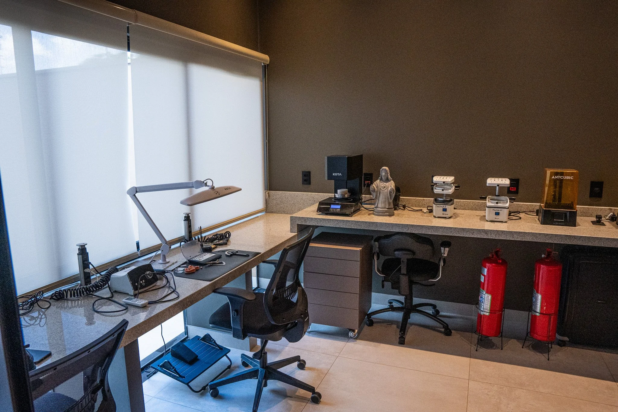 Sala de escritório com duas mesas, cadeiras, equipamentos de escritório, extintores de incêndio e diversos objetos, com janelas cobertas por persianas brancas.