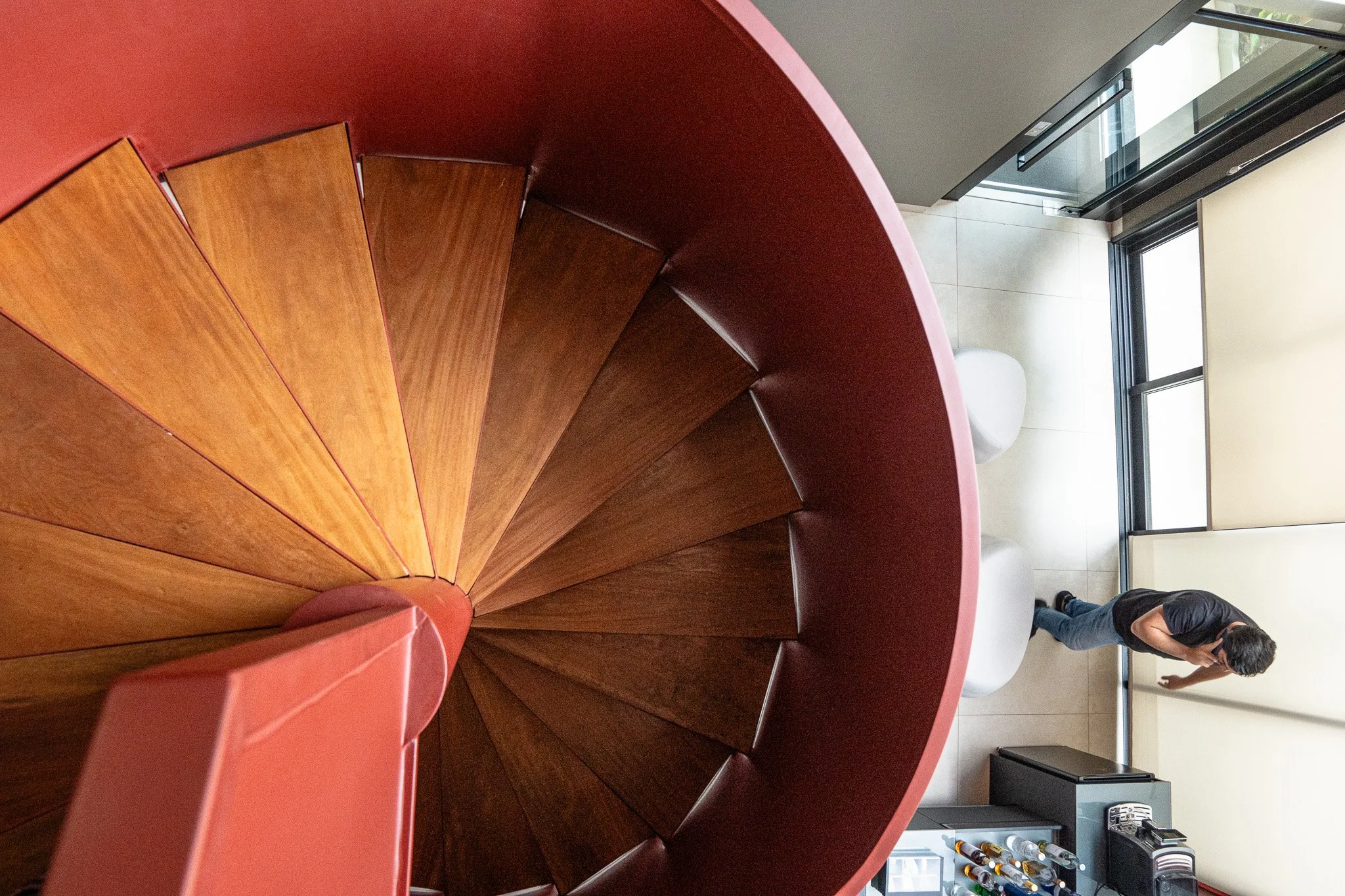Visão de cima de escada espiral de madeira com corrimão na cor vermelha, vista de dentro de um ambiente. Ao fundo, uma pessoa de camiseta preta e jeans escuros, caminhando próximo a uma porta de vidro. Há também uma área com uma mesa com garrafas e um eletrodoméstico na lateral.