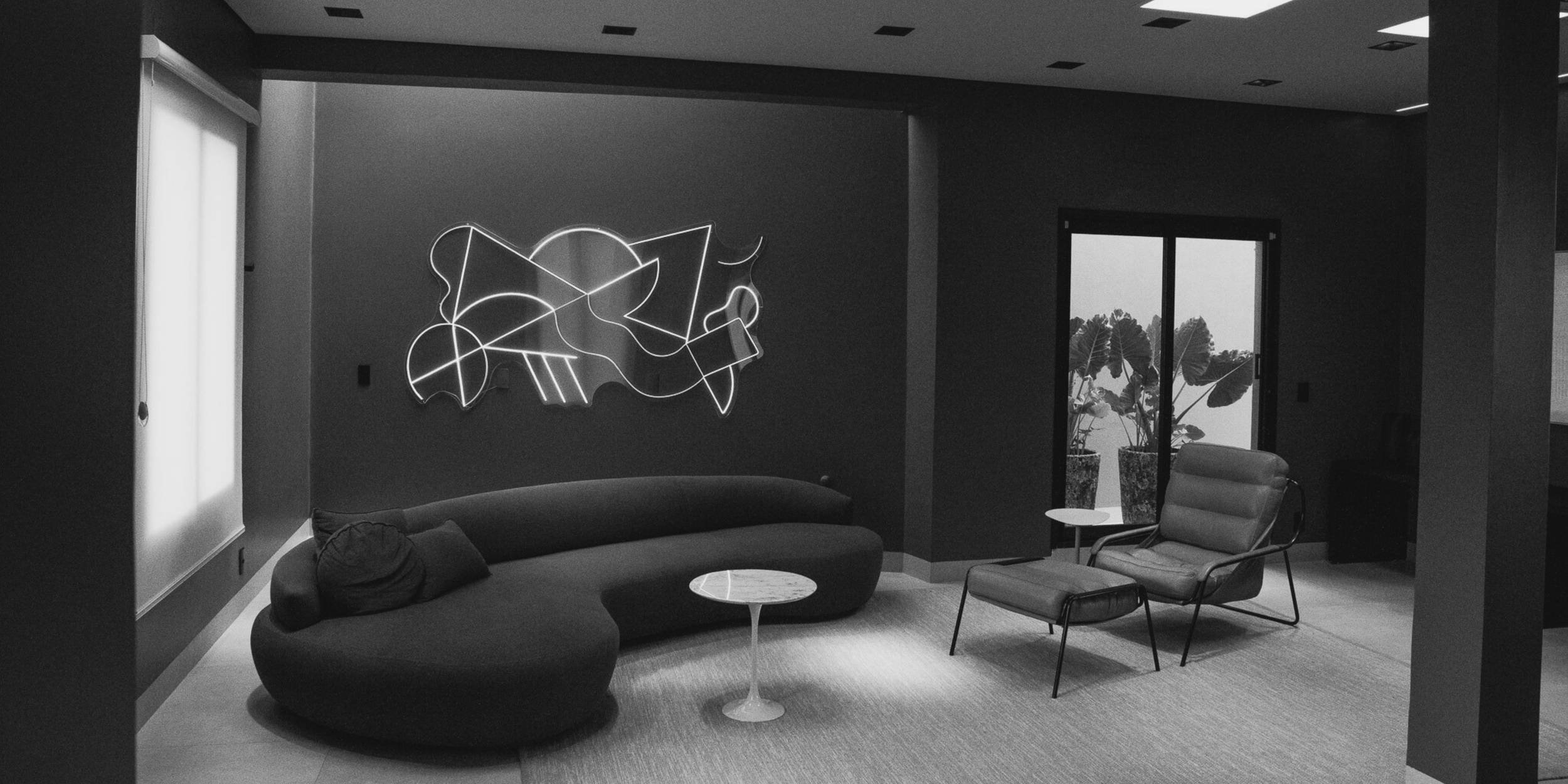 Sala de estar moderna com sofá curvo, duas poltronas, mesa de centro e iluminação de neon abstrata na parede, perto de janela com plantas.