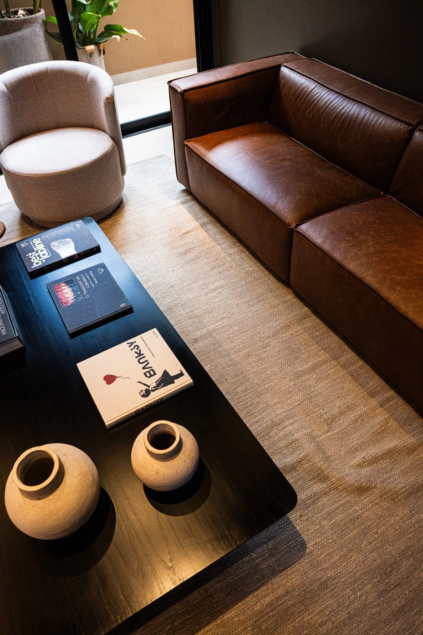 Sala de estar com uma mesa de madeira escura, duas cerâmicas decorativas, alguns livros, uma poltrona bege e um sofá de couro marrom em um ambiente de interior.