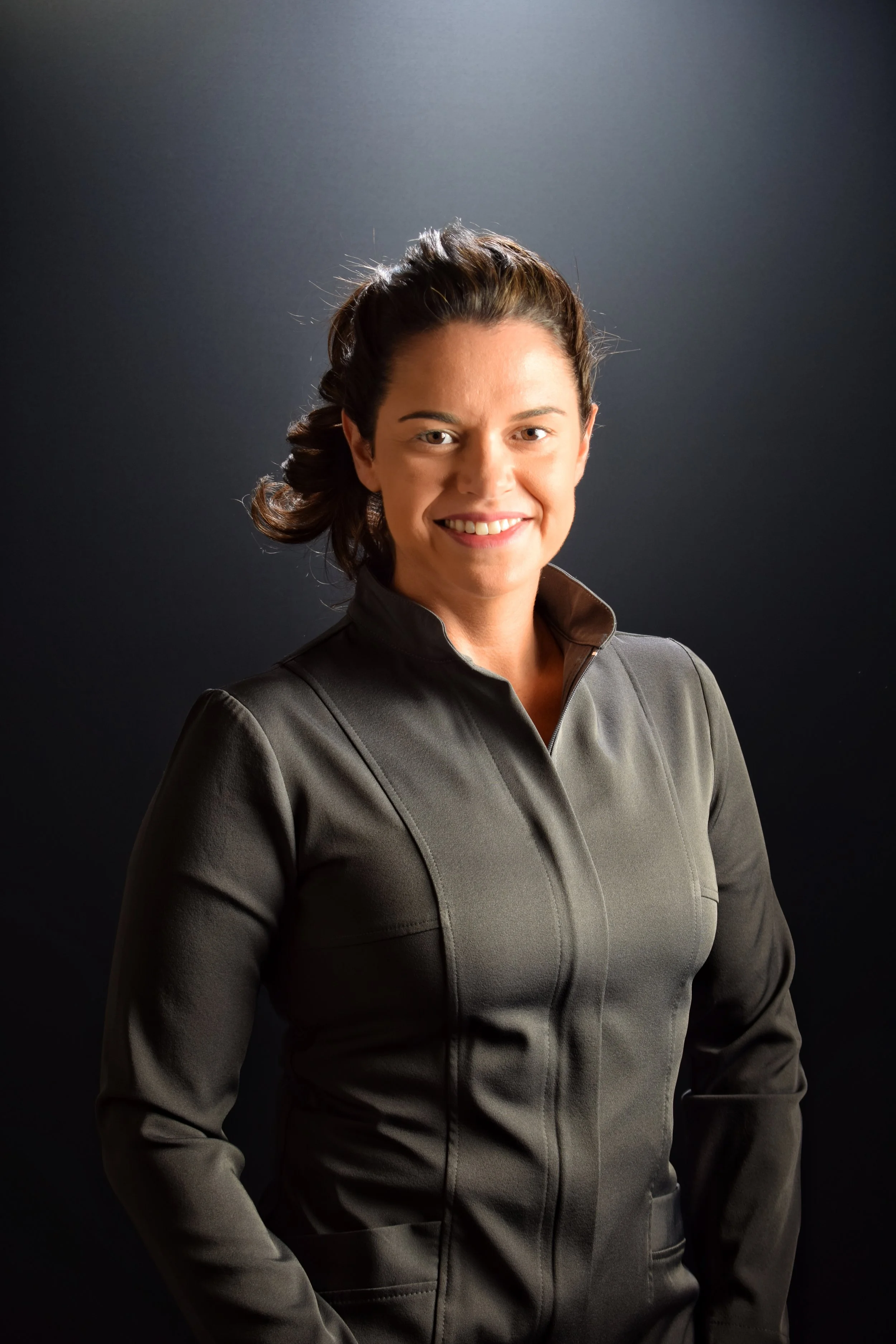 Mulher sorridente com cabelo castanho preso, vestida com jaqueta cinza, contra fundo escuro.