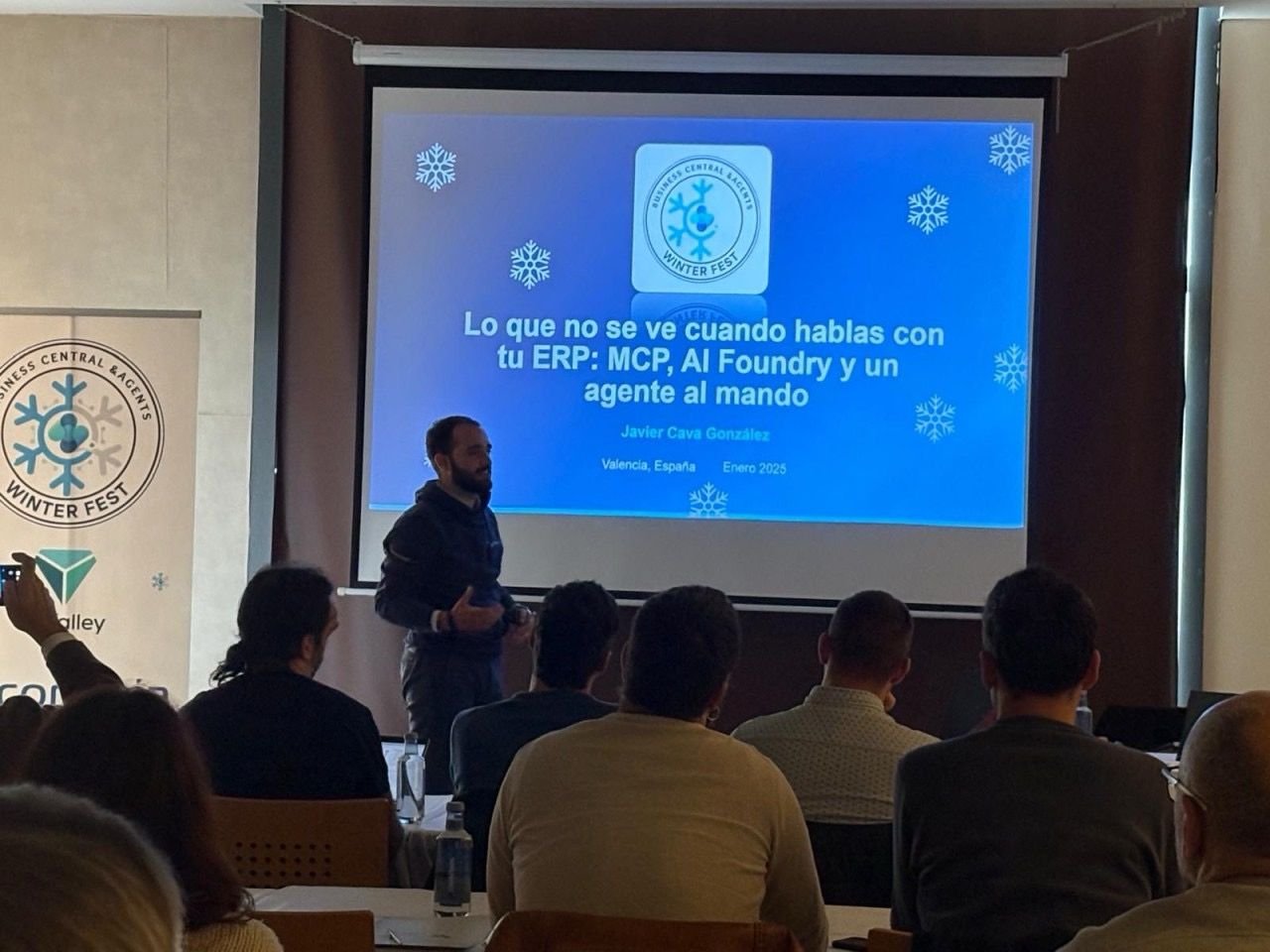 Presentador en conferencia frente a pantalla con diapositiva azul, titulada