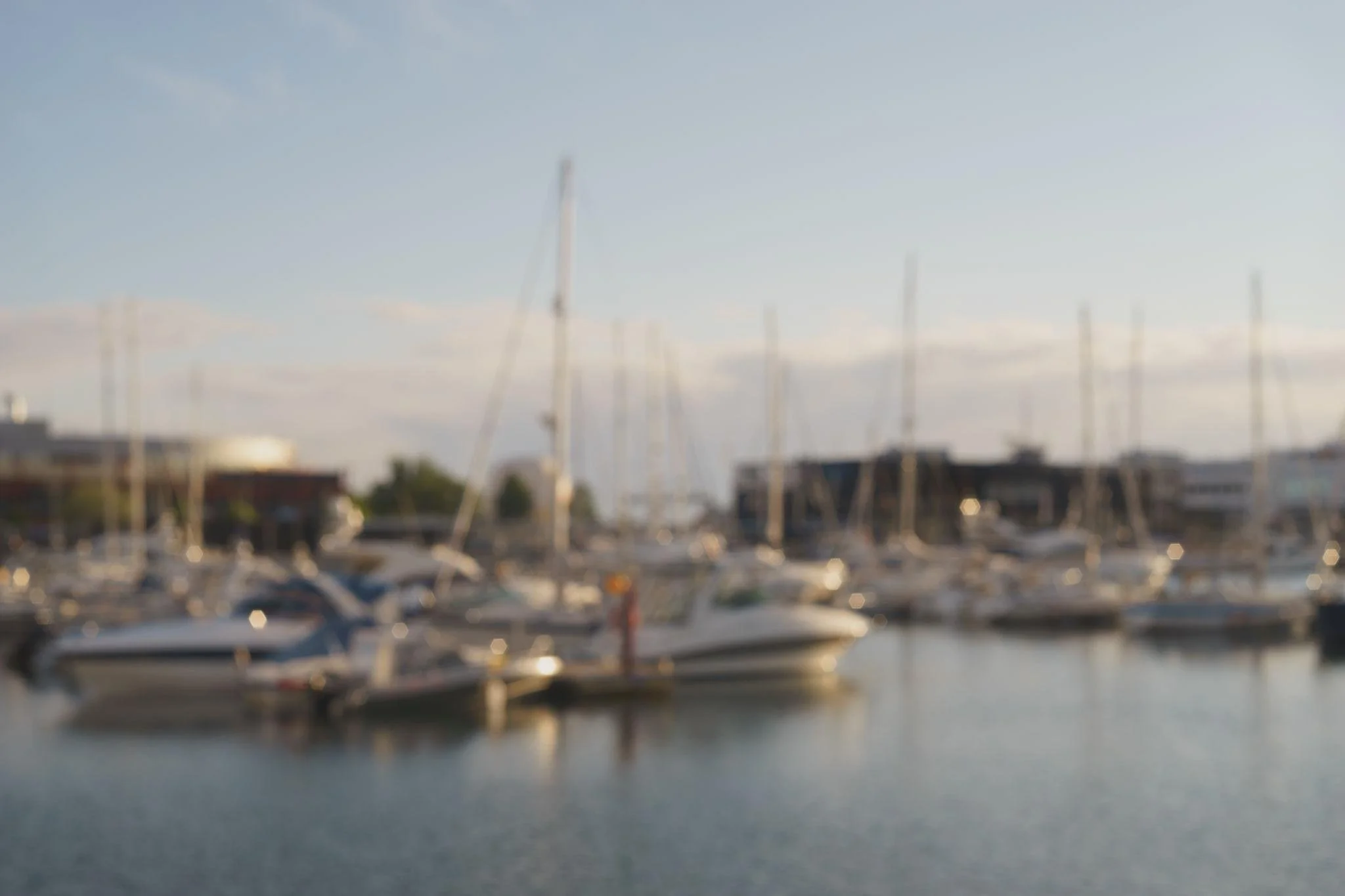 Blurry photograph of a marina with sailboats docked, buildings in the background, and a clear sky above.