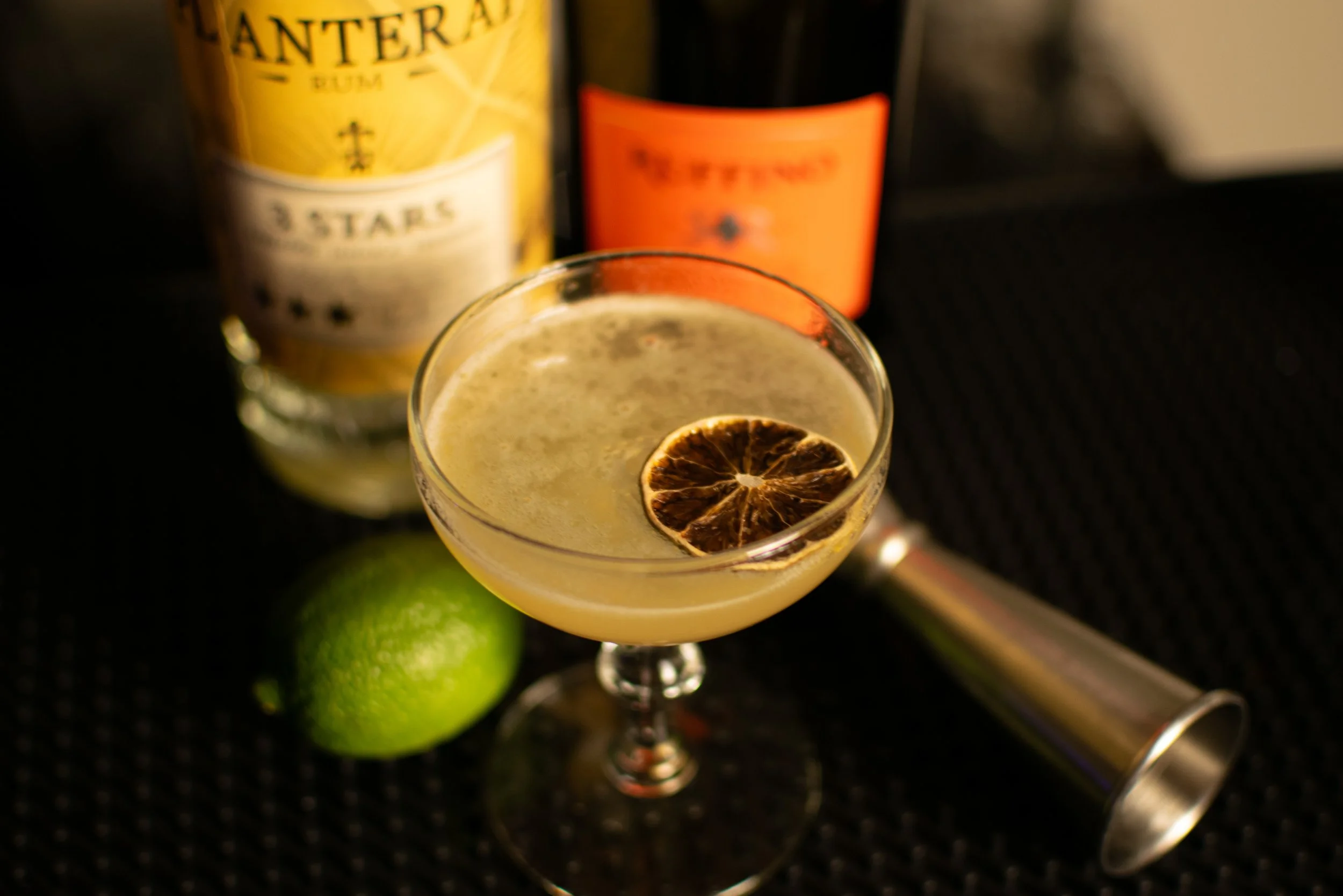 A Daiquiri cocktail with dehydrated dried lime sitting on a black surface with limes and liquor bottles in the background.