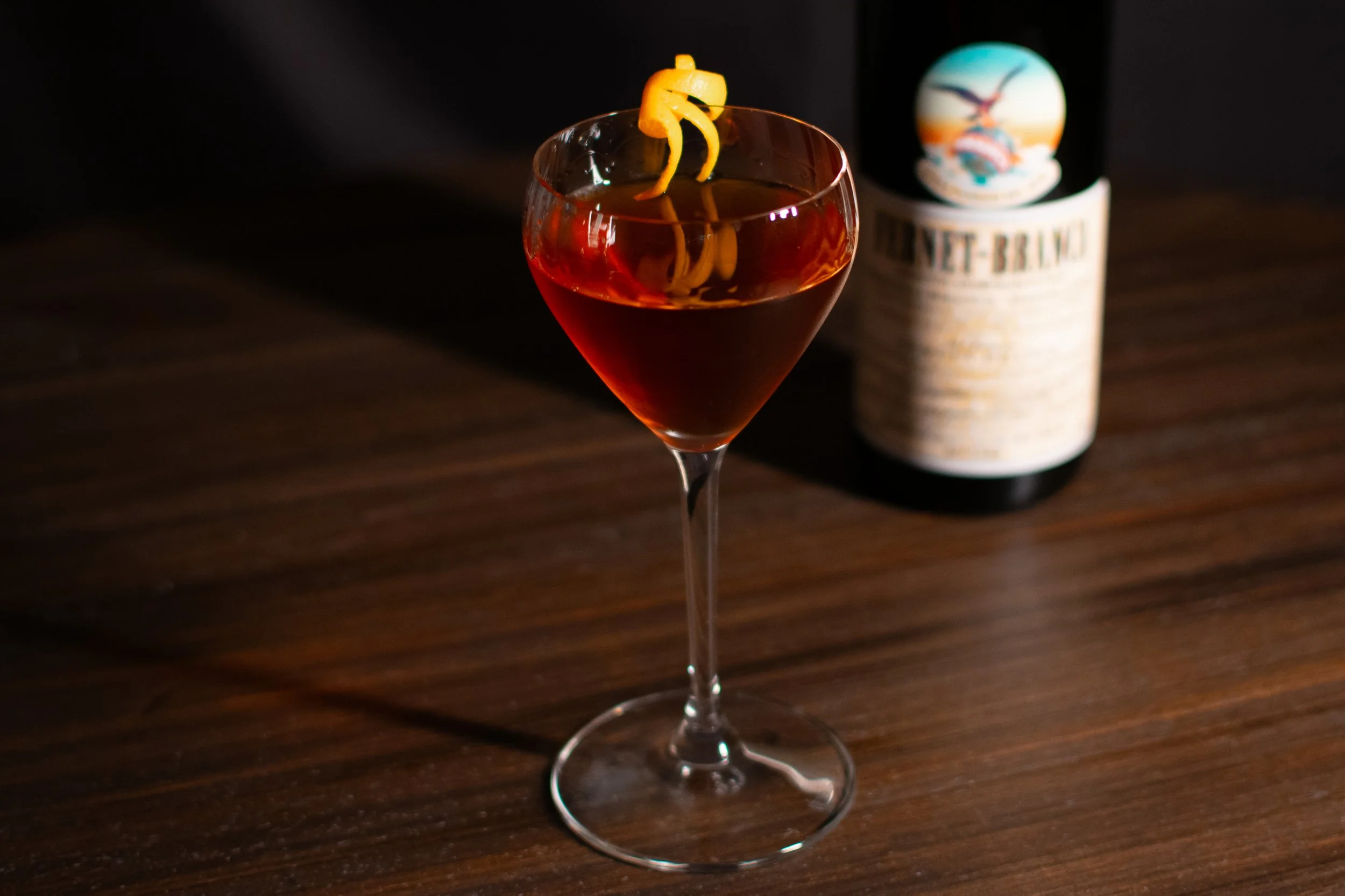 A hanky panky cocktail, garnished with a lemon twist, on a wooden surface with a bottle of Fernet Branca in the background.
