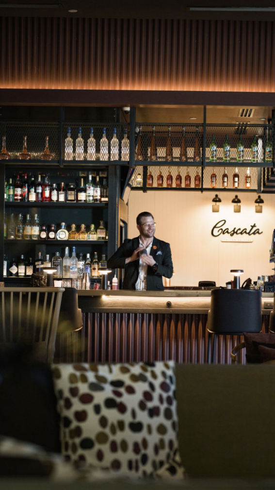 A bartender preparing a cocktail behind a bar at a restaurant named Cascata, with shelves of liquor bottles and glasses in the background.