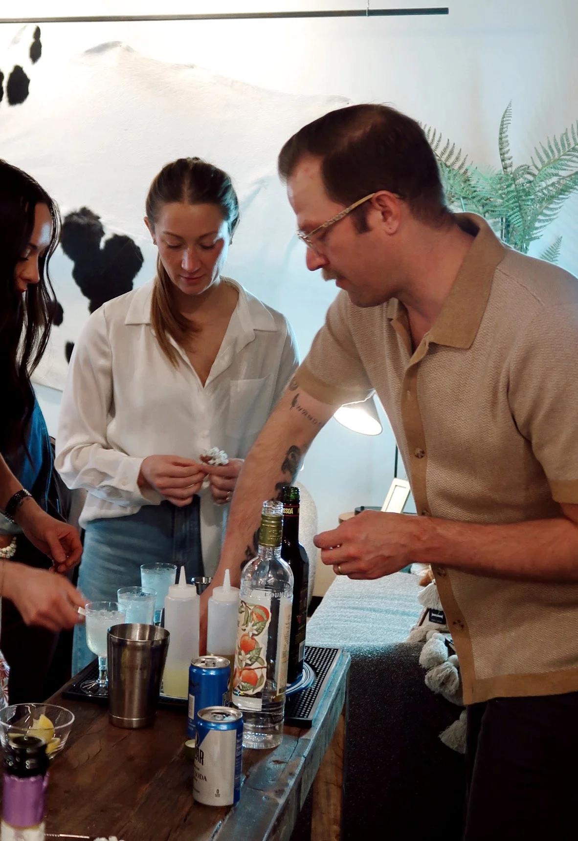 People gathered around a table with various drinks and bottles, engaged in preparing or serving beverages in a casual setting.