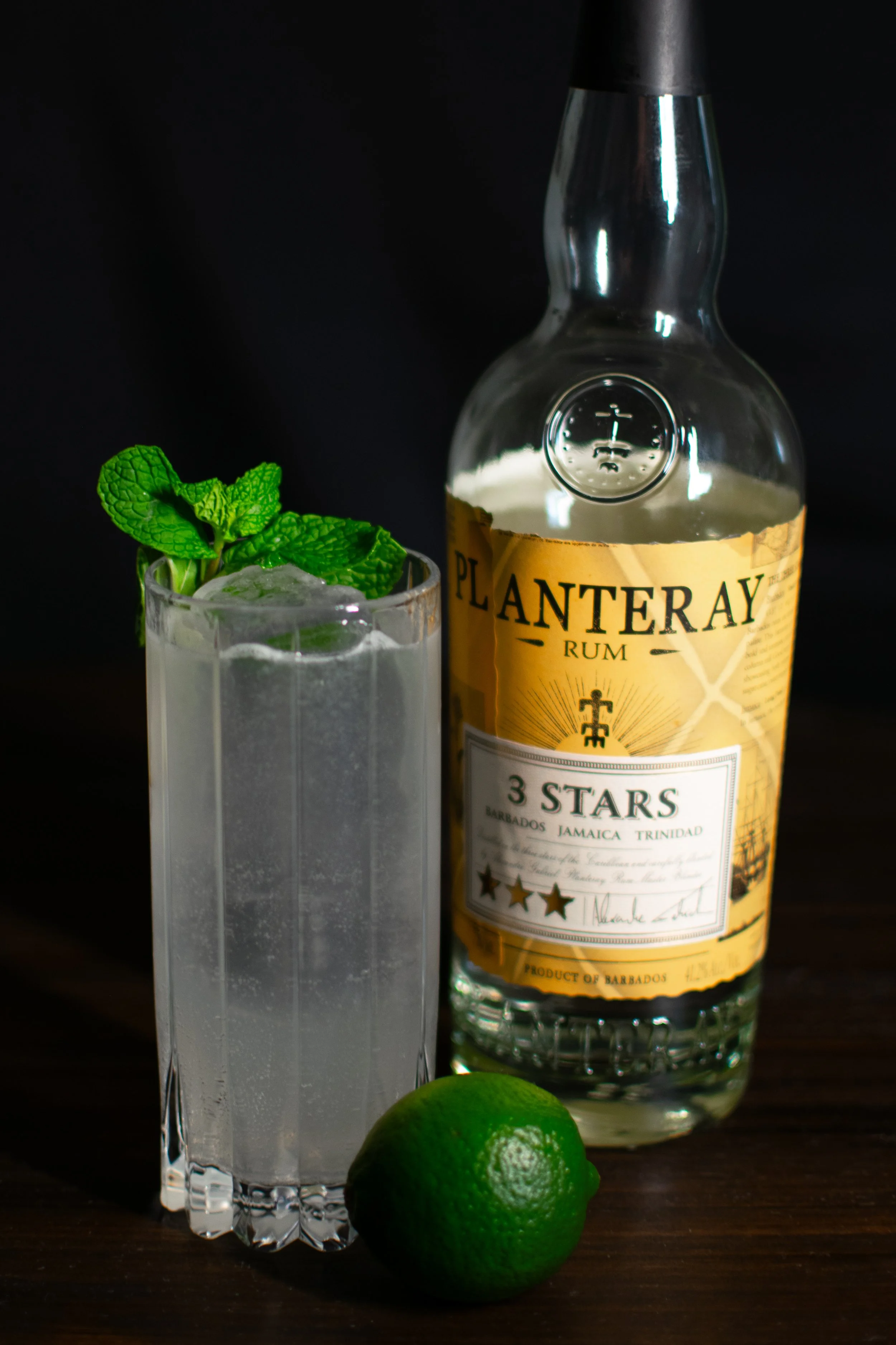 A mojito in a clear glass with a sprig of mint and ice, a bottle of Planteray Rum nearby, and a whole lime on a dark wooden surface.