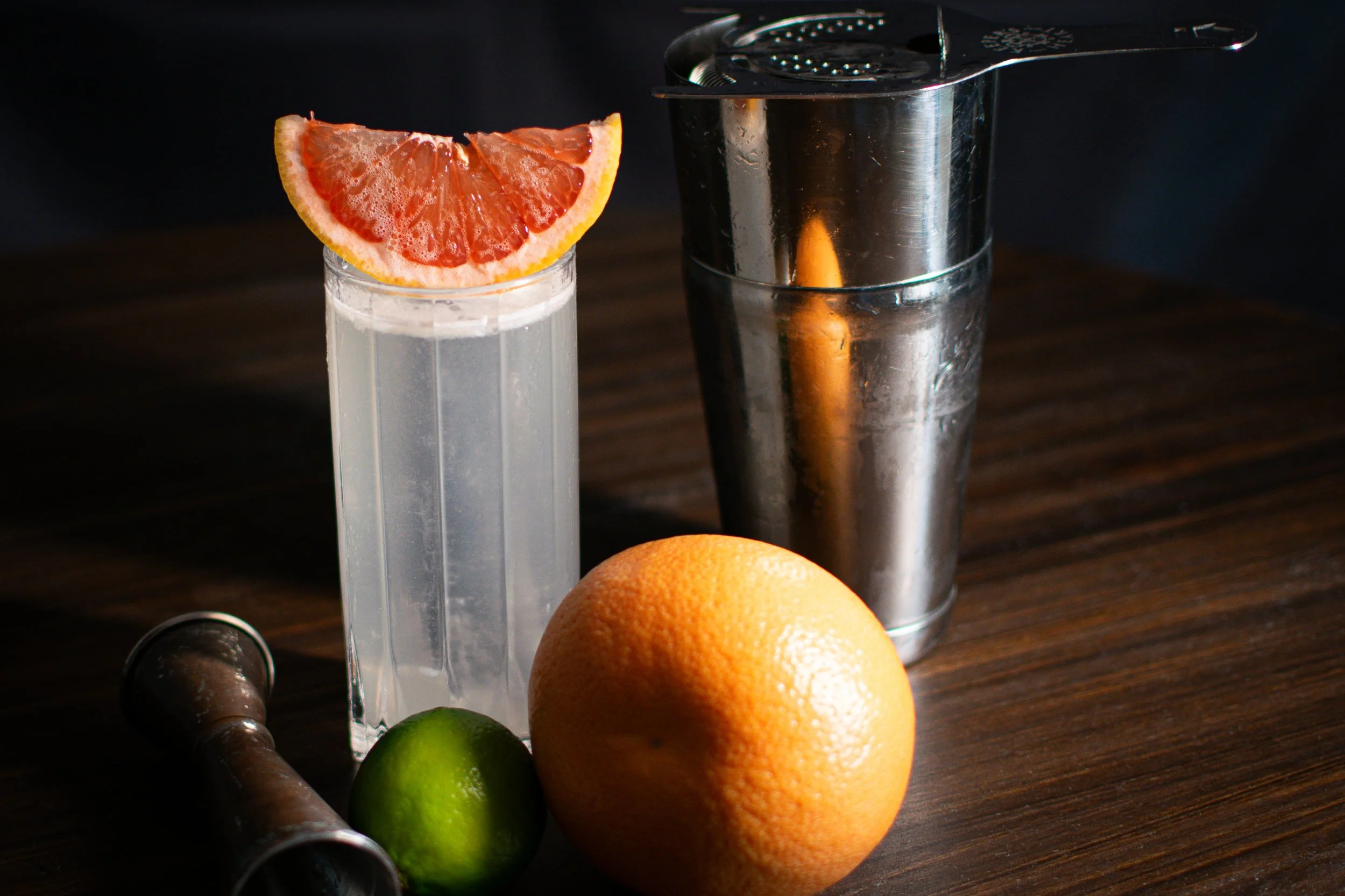 A paloma cocktail with a grapefruit wedge, a silver cocktail shaker, an orange, and a lime on a dark wooden surface.