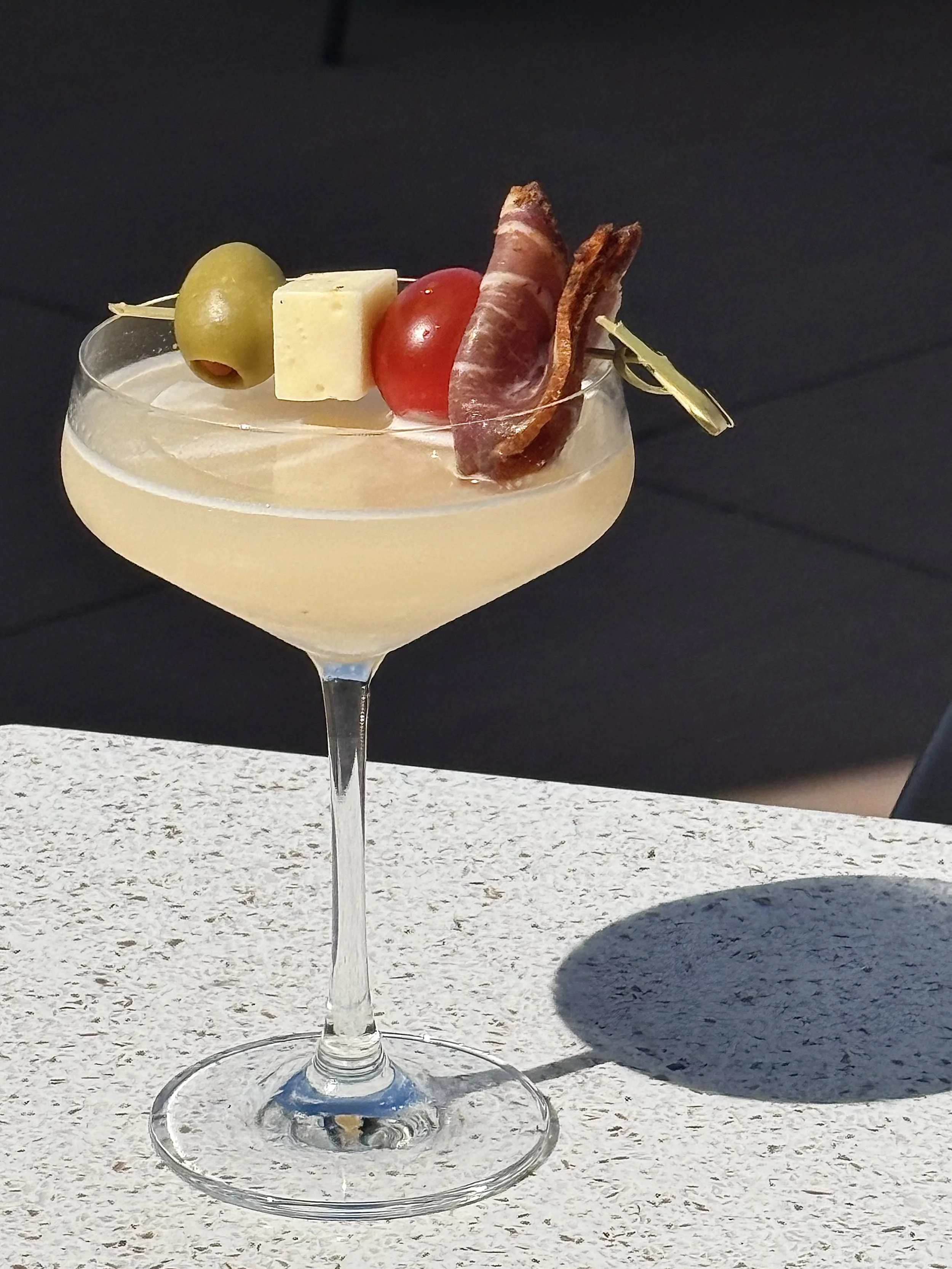 A stemmed margarita inspired by tapas cocktail glass with a yellow-colored drink, garnished with an olive, a cherry tomato, a cube of cheese, and slices of cured meat, with a black background and white textured surface.