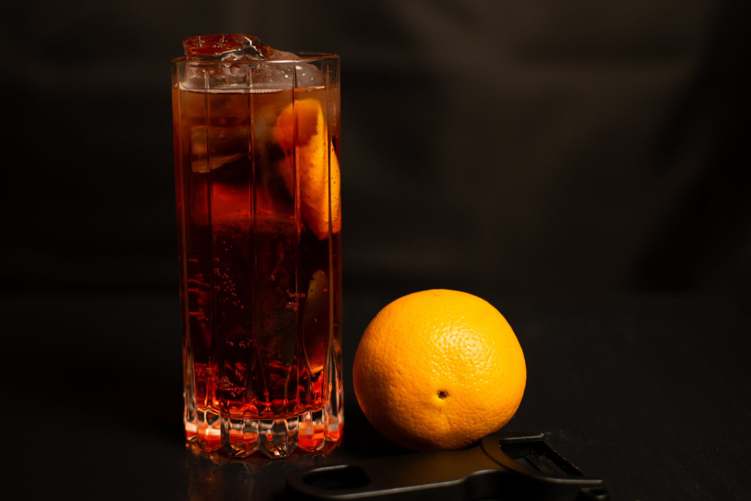 An Americano cocktail with an orange slice, placed beside a whole lemon on a dark surface.