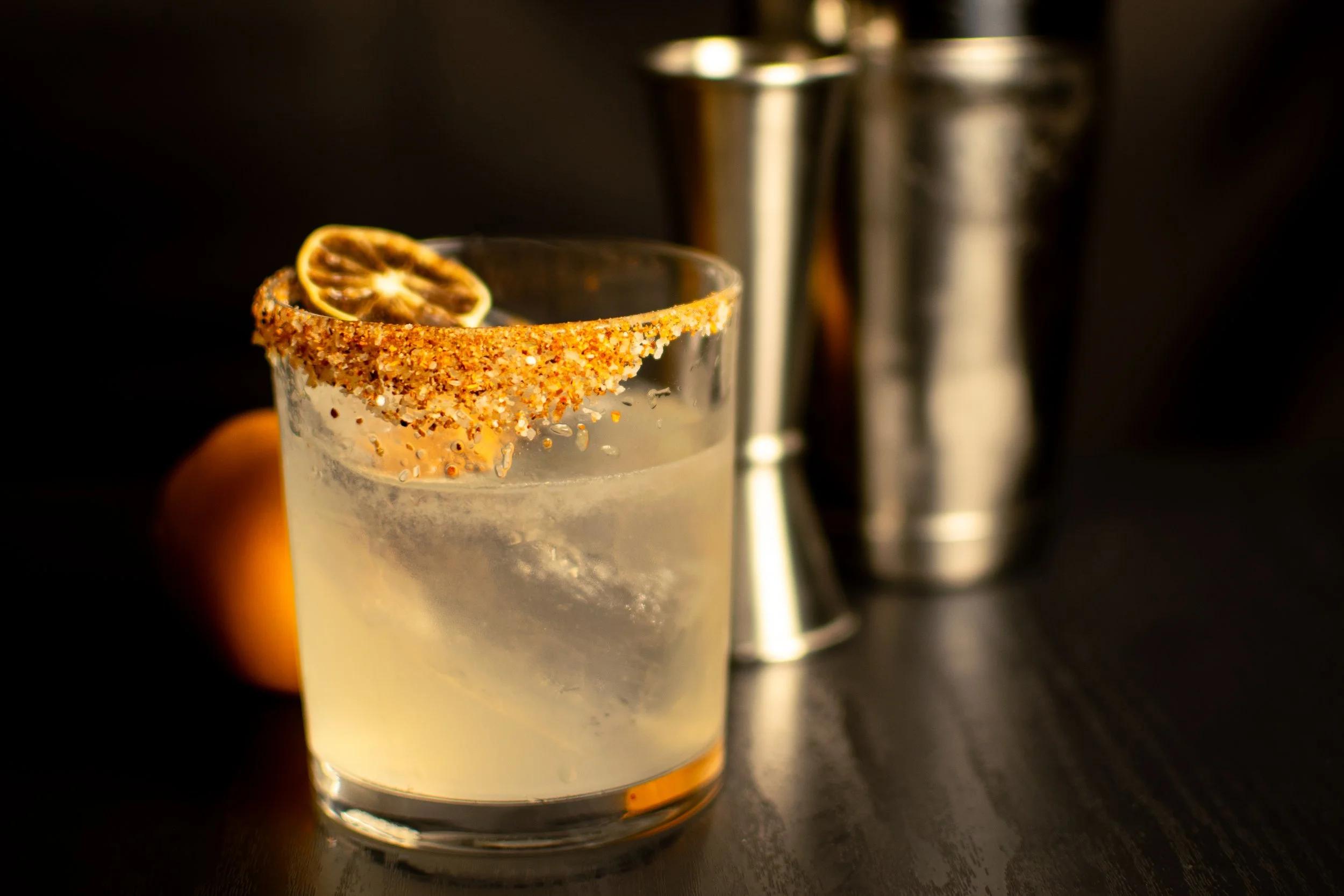 Close-up of a cocktail glass with a salted rim and a dried lemon slice garnish, with a cocktail shaker in the background.