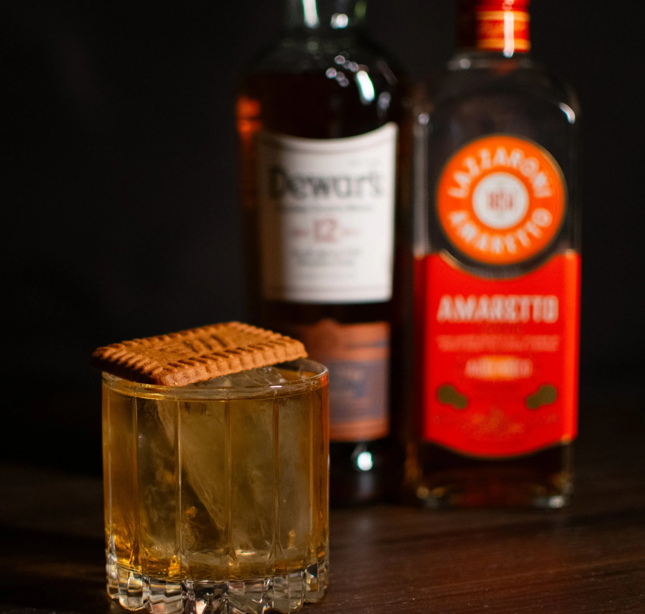 A glass of whiskey topped with a toasted graham cracker, with bottles of alcohol in the background.