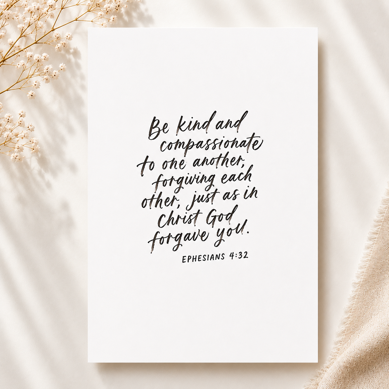 A white card with an inspirational Bible verse from Ephesians 4:32 written in black cursive font, surrounded by light beige fabric and delicate white flowers.