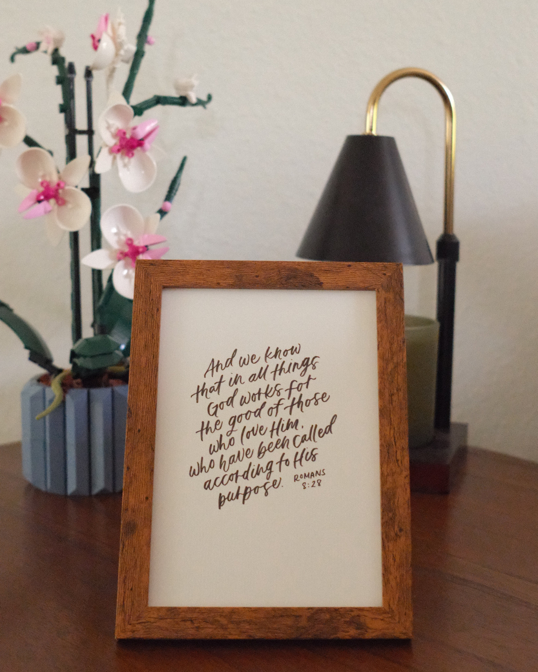 A framed handwritten quote on a table, with a potted plant with white and pink flowers and a black and gold desk lamp in the background.