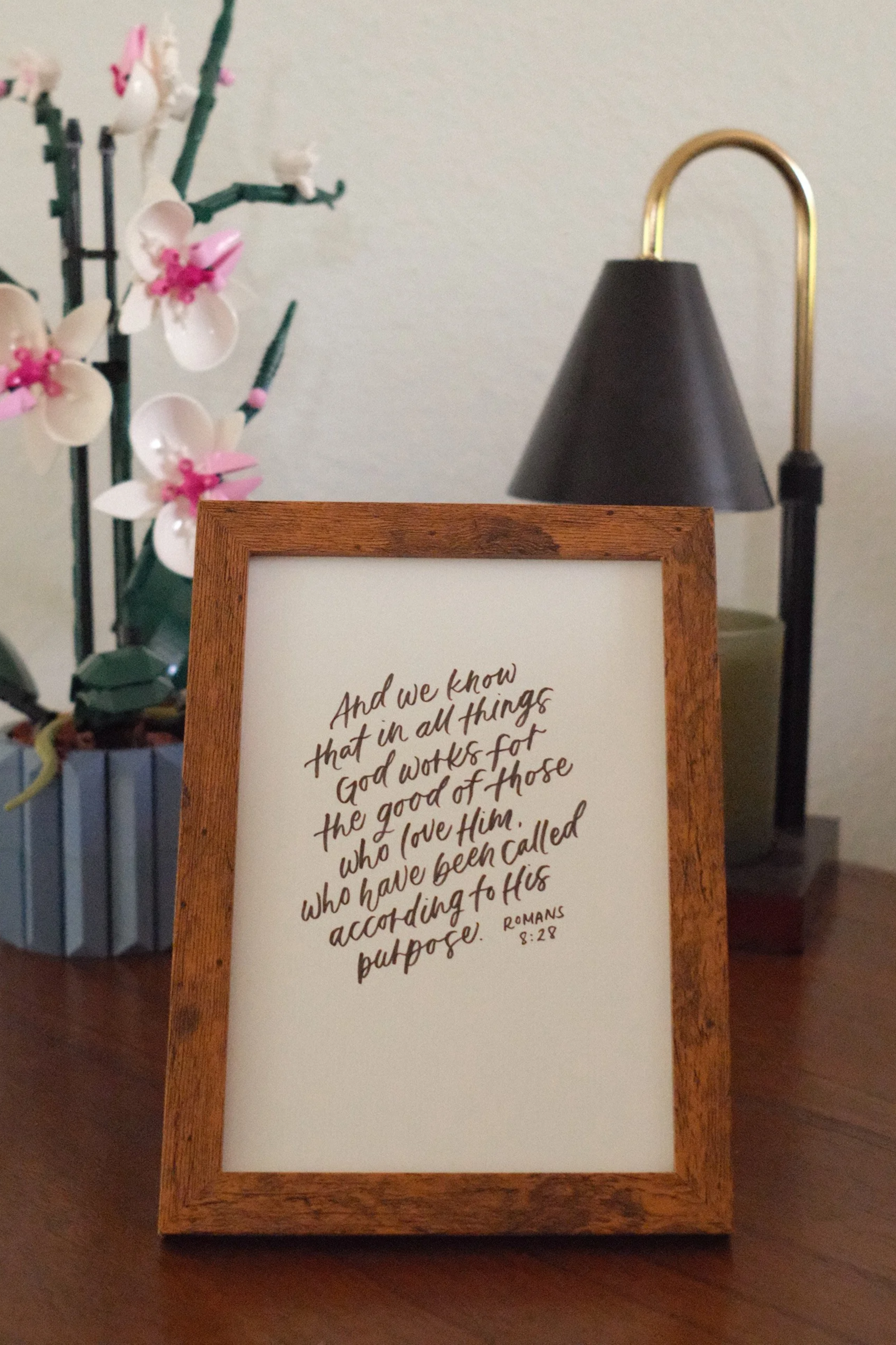 A wooden framed sign with a quote from Romans 8:28 written in cursive at the bottom, placed on a wooden surface. Behind it, there is a black lamp with a gold arm and a flower arrangement with white and pink flowers.