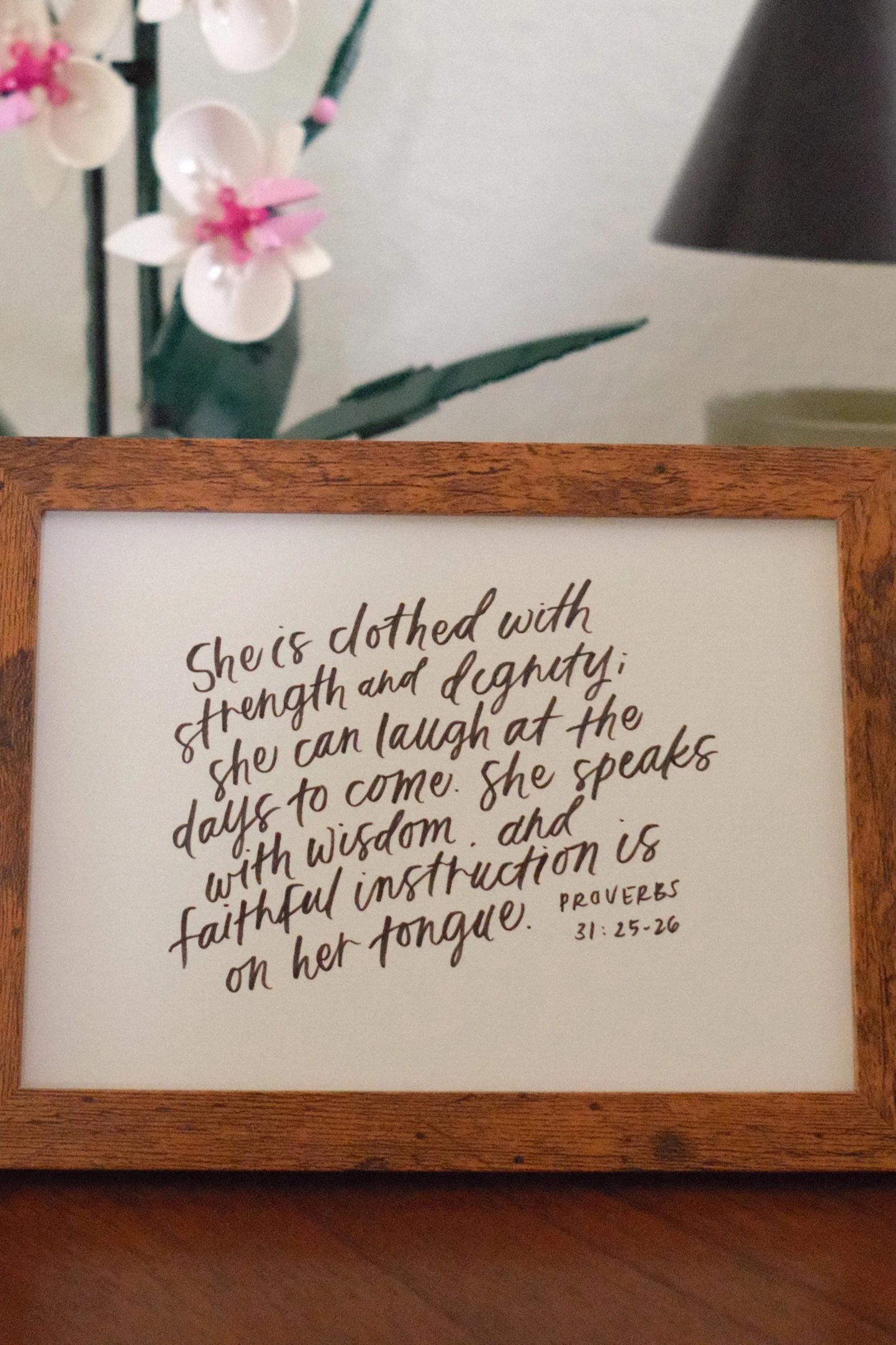 A handwritten quote on a white paper framed in wood, with part of a flower arrangement and a black lamp visible in the background.