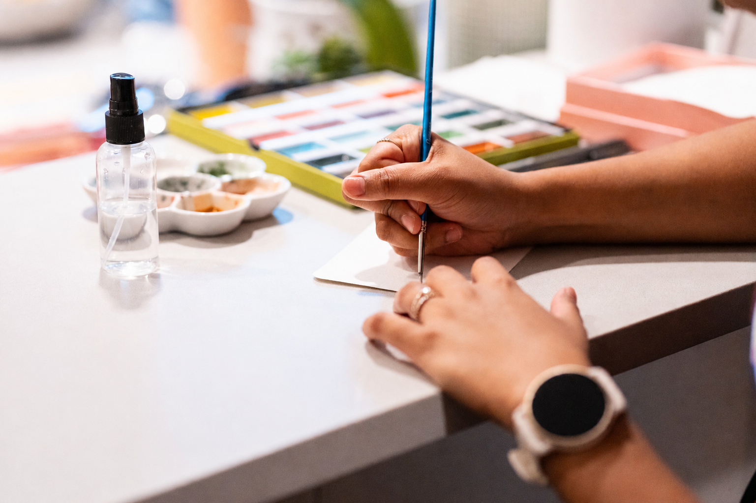 A person is painting with watercolors at a desk, with a palette, a spray bottle, and watercolor set in the background.