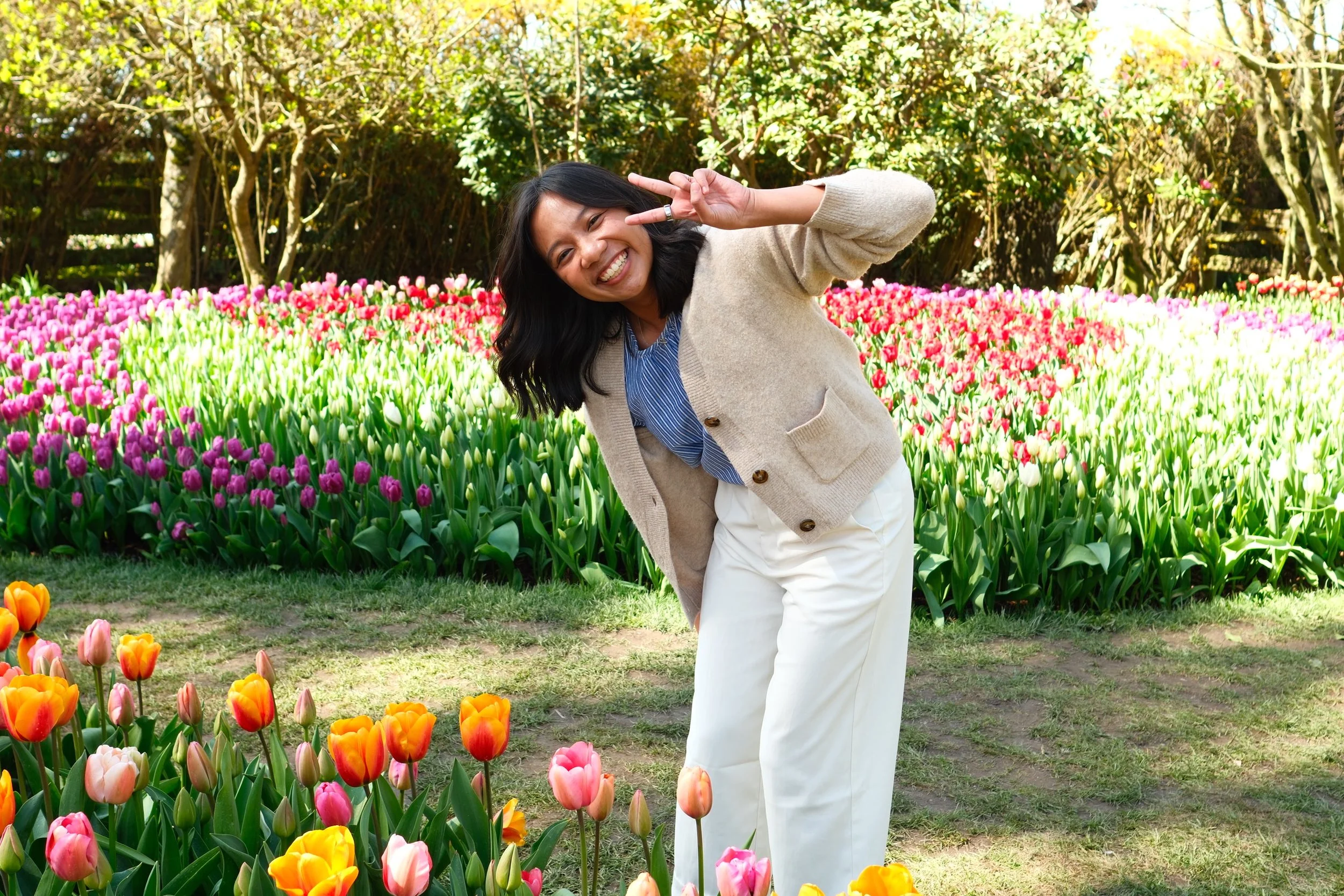 A woman smiling and making a peace sign in a garden full of colorful tulips.