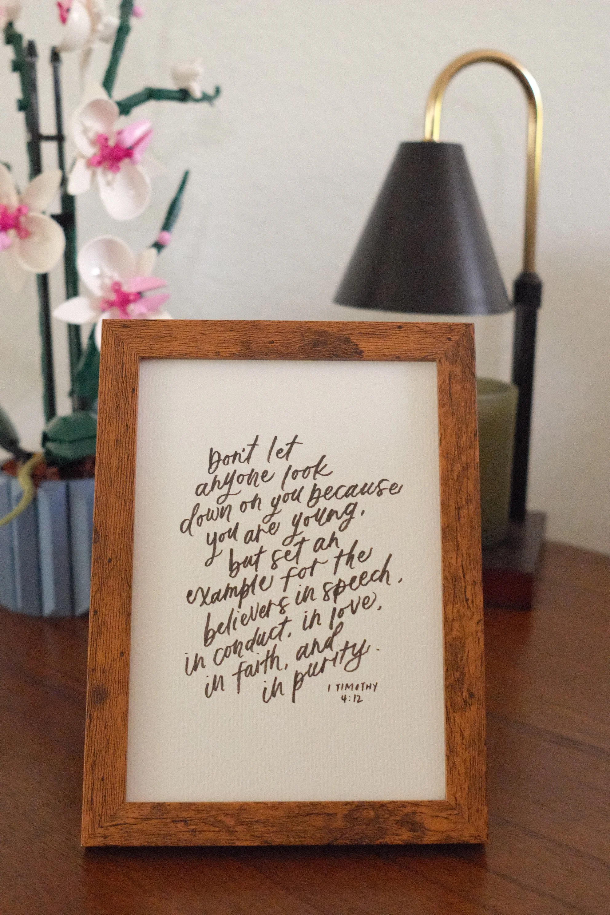 A framed handwritten quote on a piece of paper, placed on a wooden stand. In the background, there is a black lampshade with a brass curved support and a blurred image of a plant with white and pink flowers.