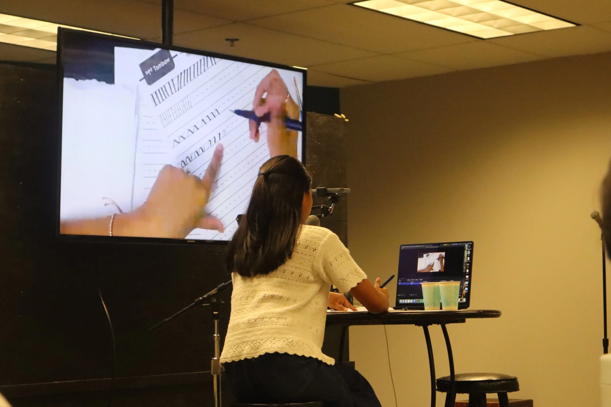 A woman with long dark hair, dressed in a yellow sweater, is sitting at a table with a laptop, participating in a presentation or workshop. A large screen displays her writing on lined paper with a pen. The setting appears to be an indoor conference or seminar room with yellow walls and ceiling lights.