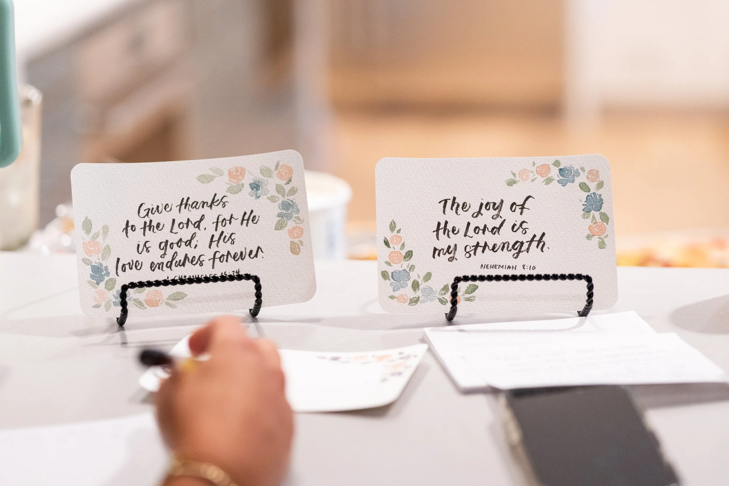 Close-up of a hand writing on a card with floral border, with two other cards with similar floral borders and religious texts in the background.