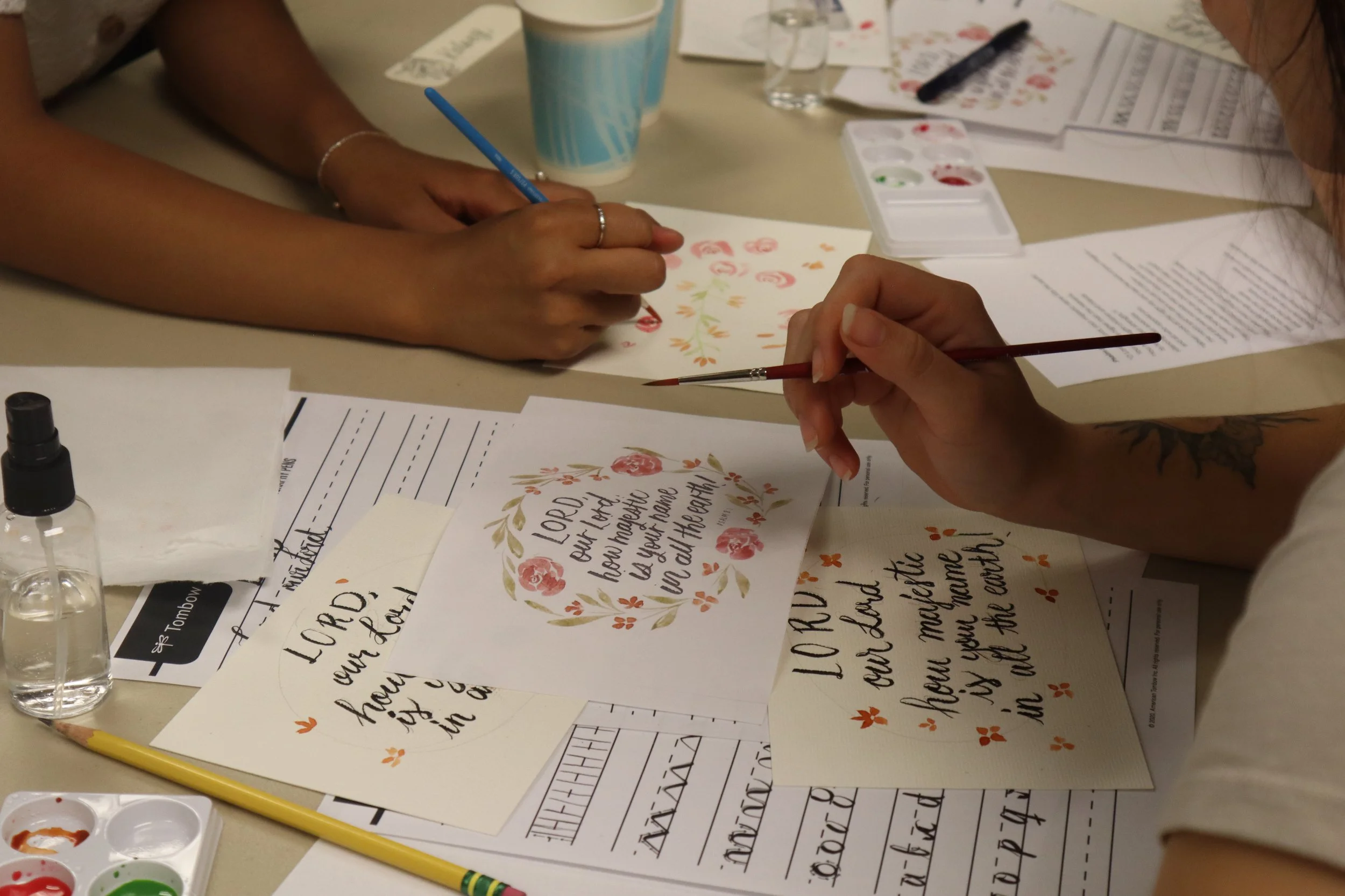 Close-up of three hands working on decorative calligraphy and watercolor projects on a table scattered with papers, watercolor paints, and pens.