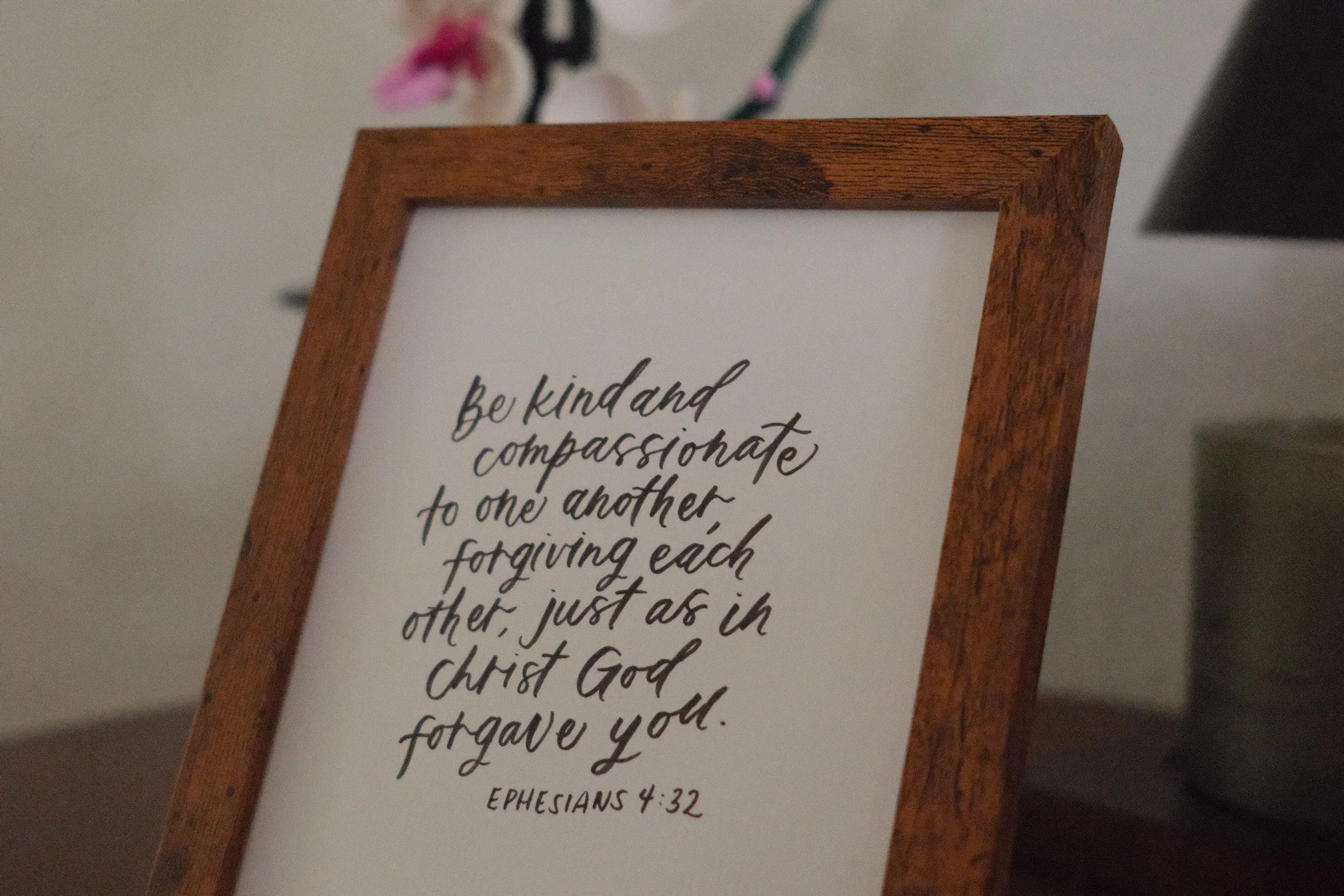 Close-up of a framed printed quote on a white background with handwritten script, placed on a flat surface. The quote reads, 'be kind and compassionate to one another, forgiving each other, just as in Christ God forgave you. Ephesians 4:32'.