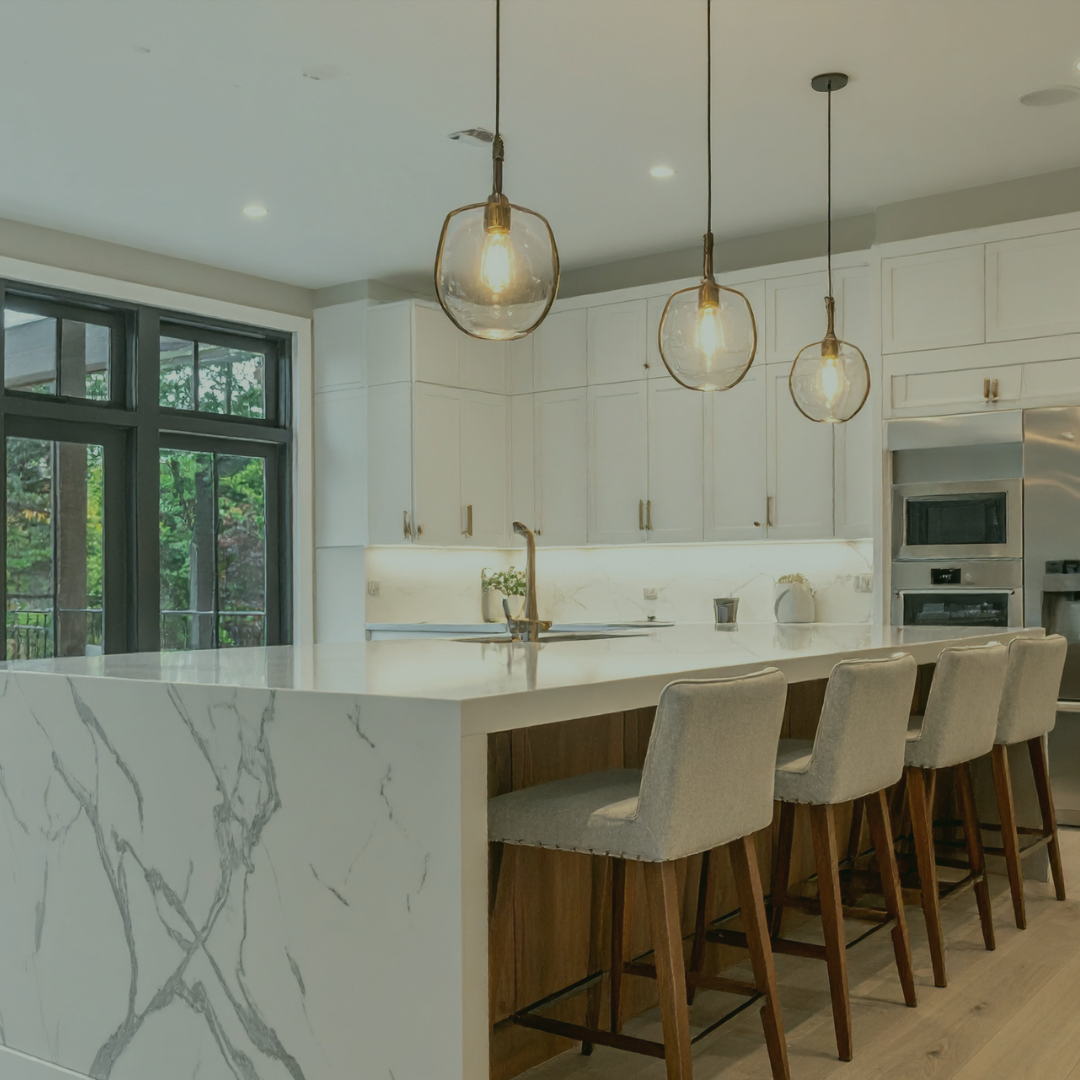 Modern kitchen with white cabinets, marble island, three pendant lights, large window showing greenery, and beige chairs.