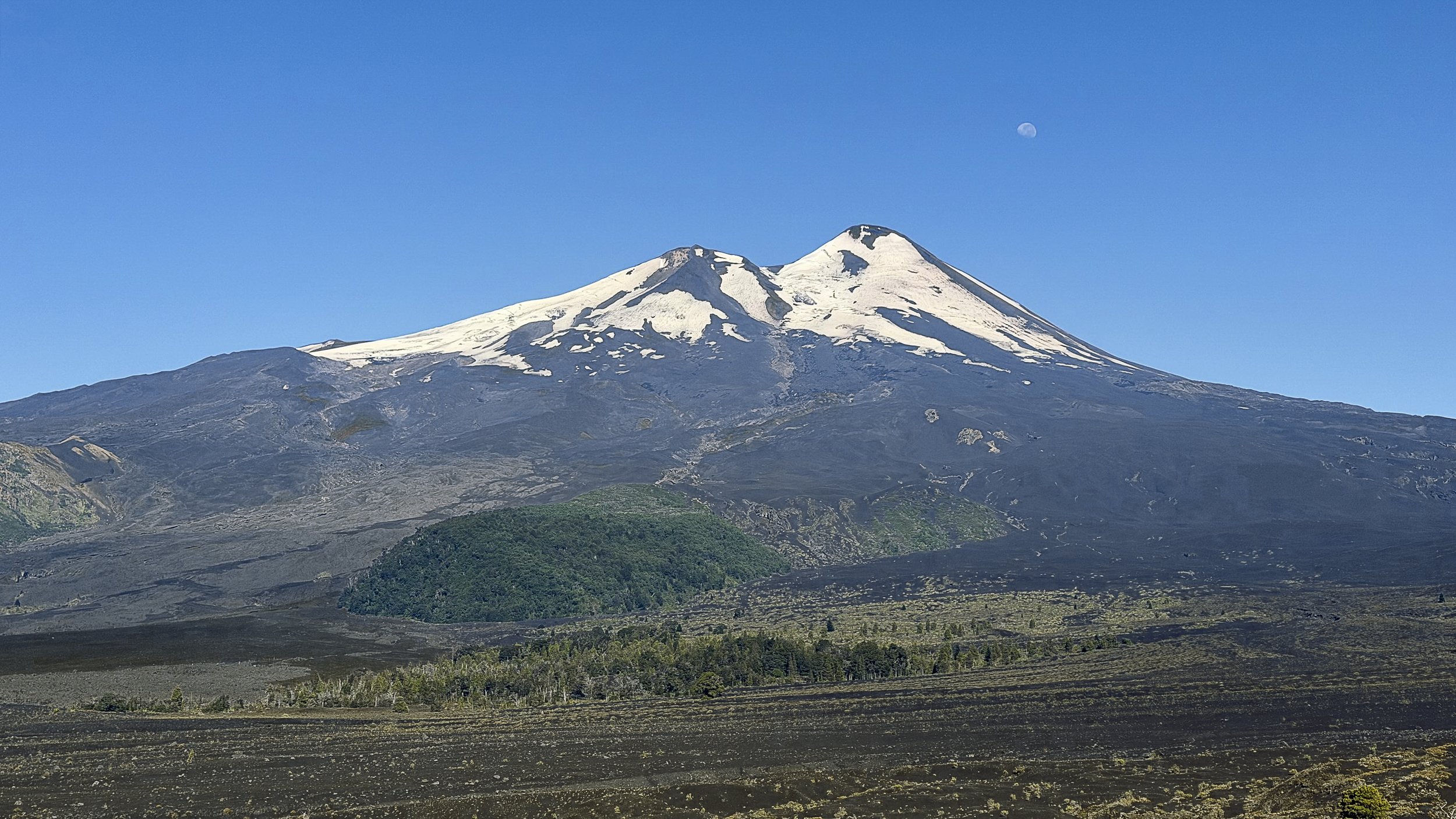 Un grand volcan avec deux sommets enneigés, sous un ciel clair bleu avec la lune visible en haut à droite.