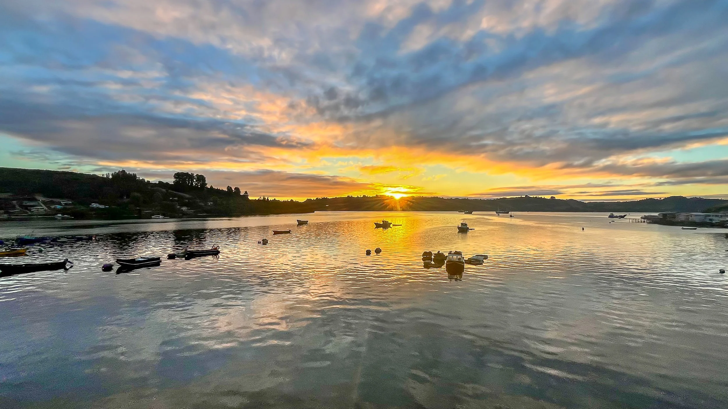 Coucher de soleil sur une rivière avec plusieurs bateaux flottant à la surface, des maisons en arrière-plan et un ciel nuageux.