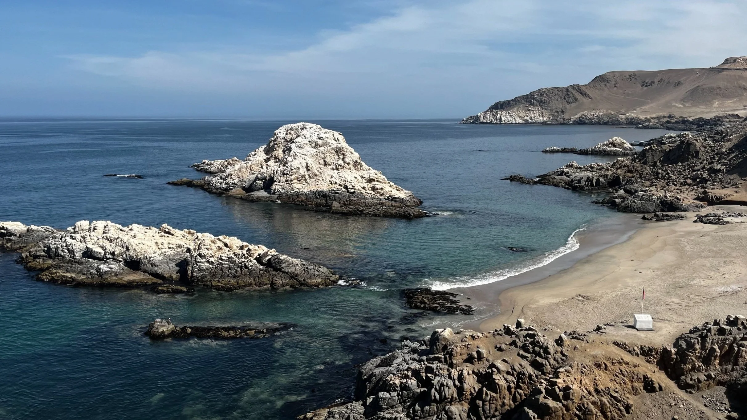 Plage rocheuse avec un rocher blanc entouré d'eau bleue et une plage de sable clair, avec des collines en arrière-plan.
