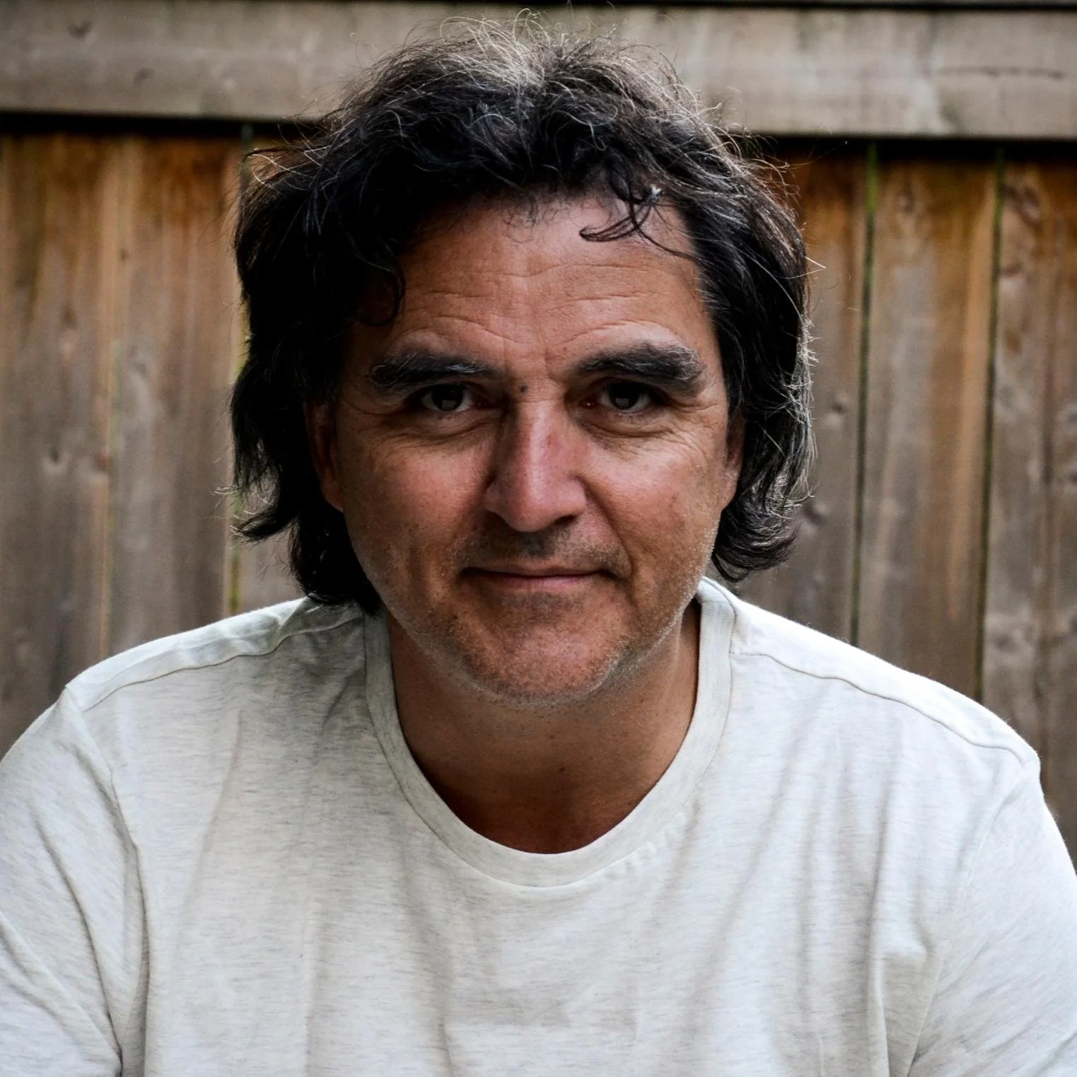 Un homme avec des cheveux noirs, portant un t-shirt blanc, posé devant une clôture en bois dans un jardin.