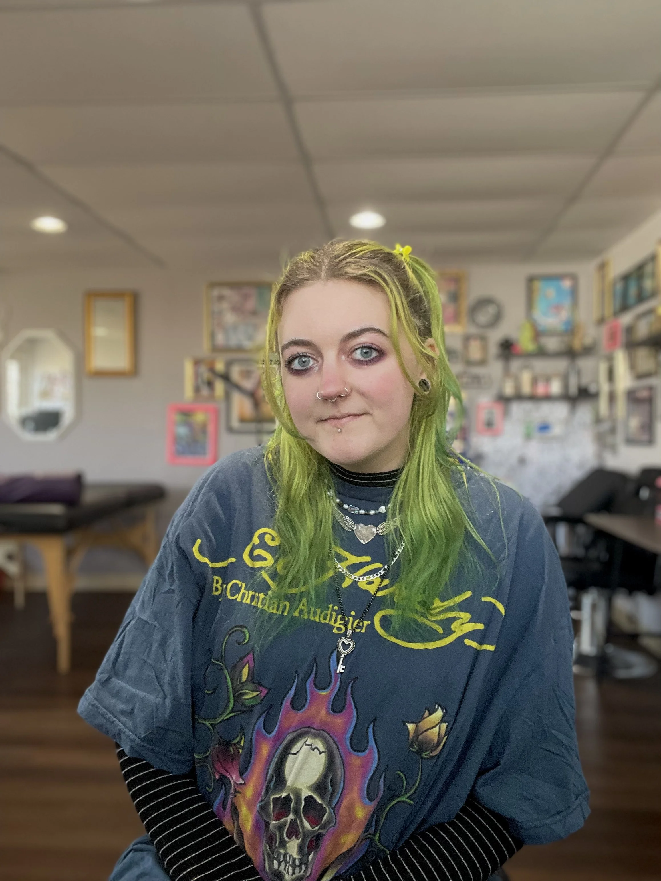 Young woman with green hair, wearing a gray T-shirt with a skull and flames design, sitting in a room with framed pictures and books on the walls.
