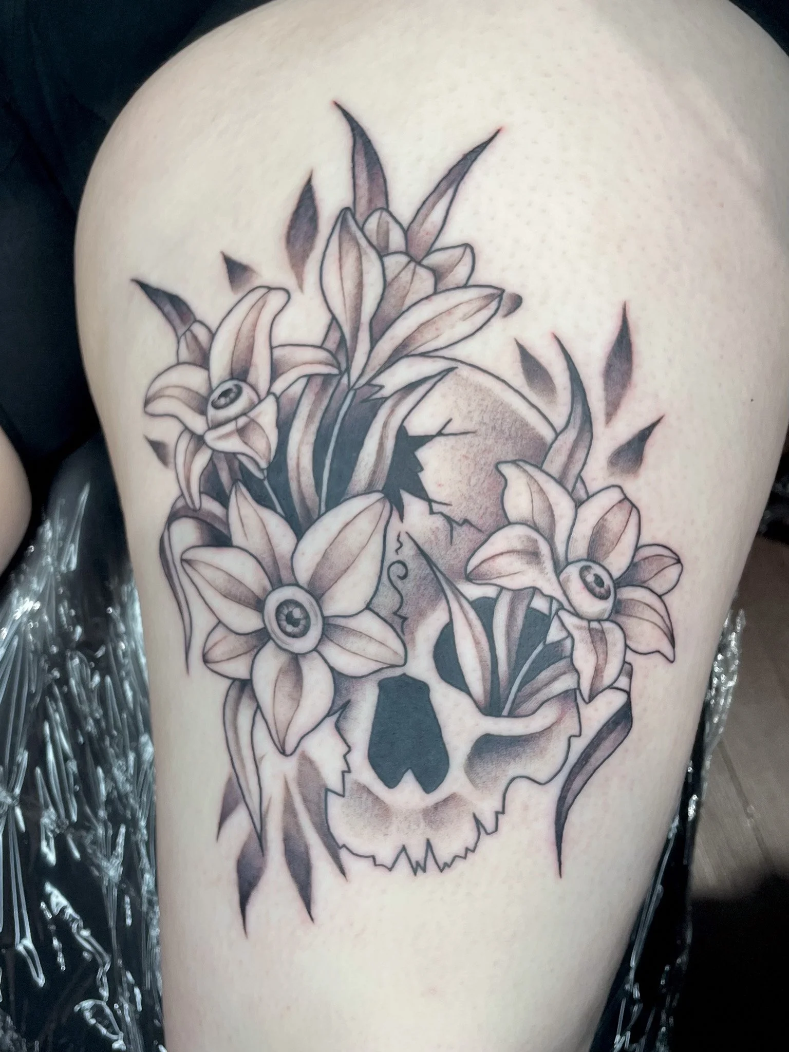 Tattoo of a skull with lilies and an eye within the flowers on a person's thigh.