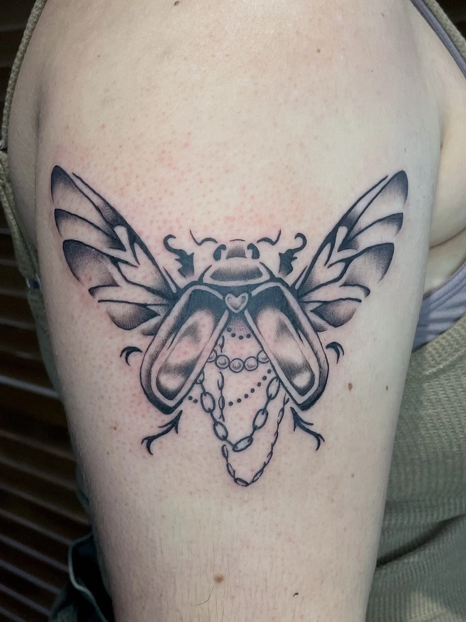 Tattoo of a bee with wings, a heart shape, and chain details on a person's upper arm.