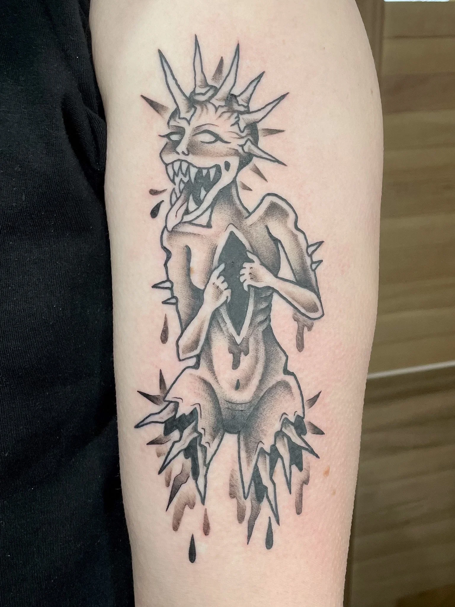 Black and gray tattoo of a monstrous creature with a snout, sharp teeth, and spiky head, holding a diamond-shaped object with dark liquid dripping from it.