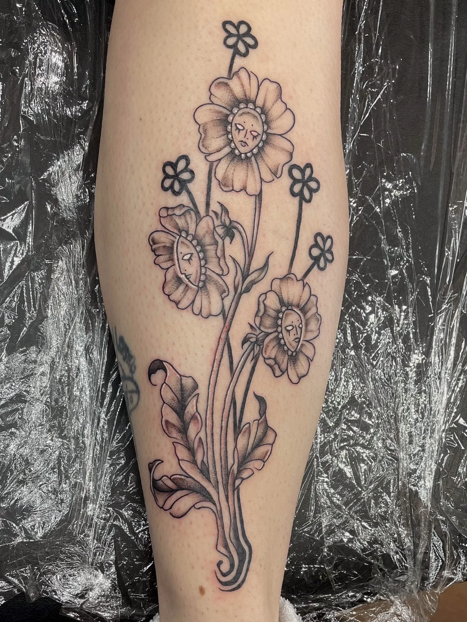 Black and gray floral tattoo with five flowers, each with a face inside the petals, on a person's arm.
