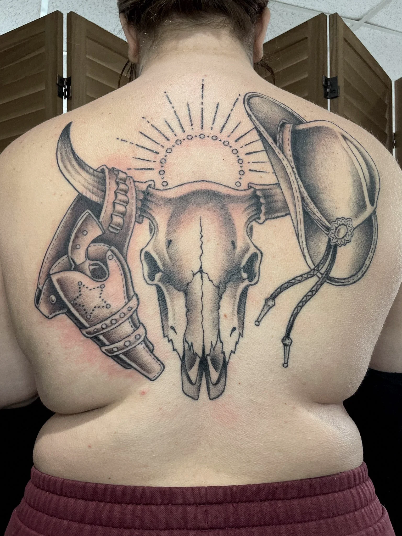 Tattoo of a cow skull with a cowboy hat, sheriff badge, and a sunburst design on the back.