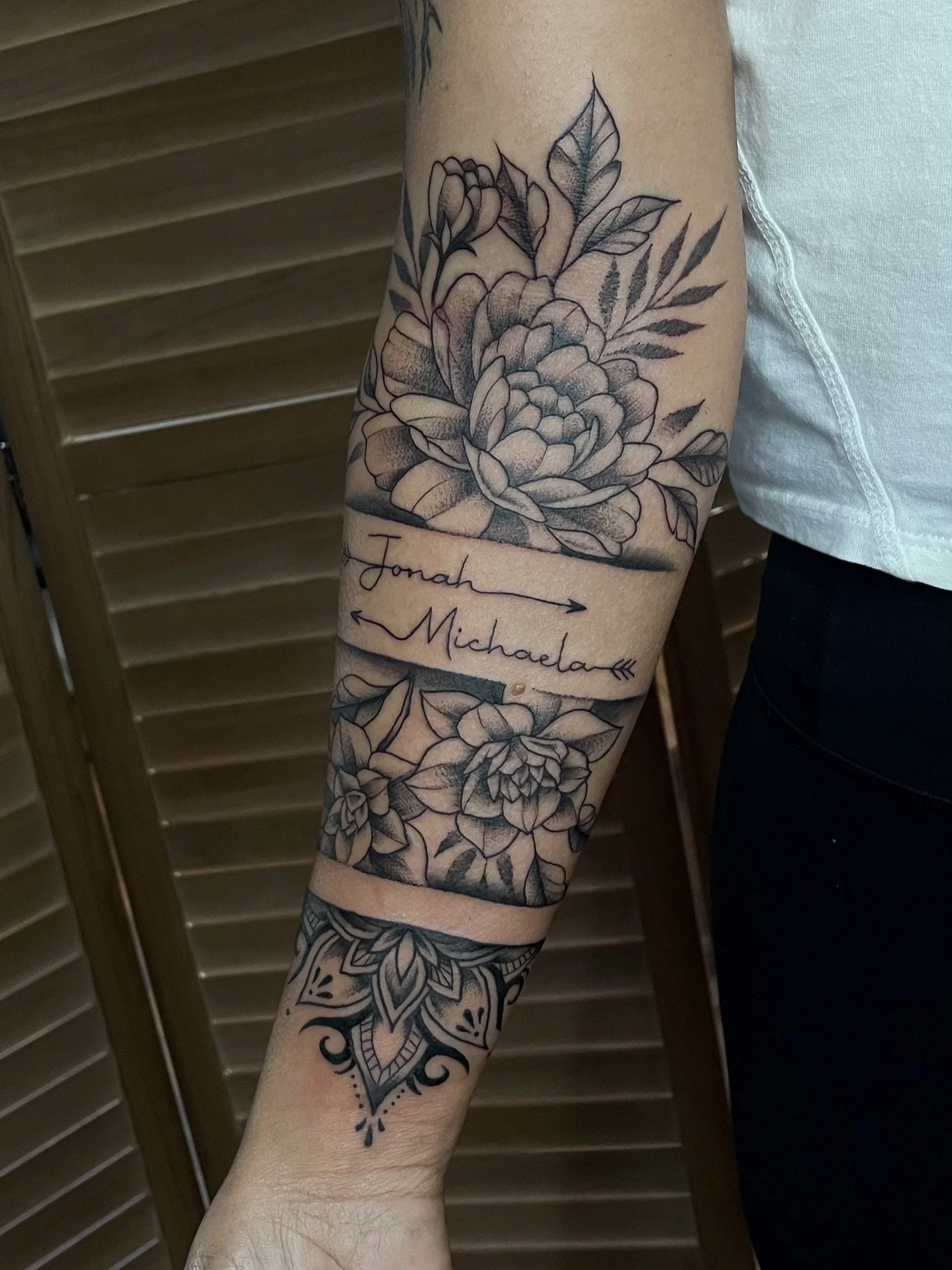 Black and gray floral tattoo on person's inner forearm with two large flowers, leaves, and a banner with the names 'Jonah' and 'Michaela' connected with arrows.