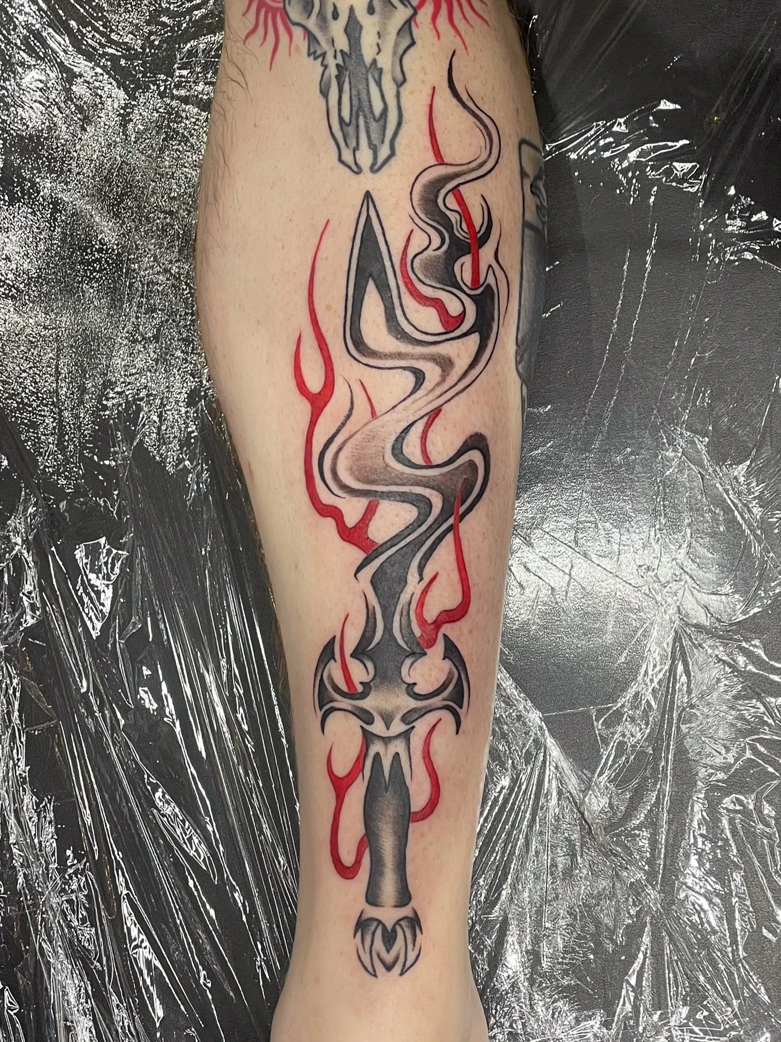 Tattoo of a dagger with a tribal design, surrounded by red flames, on a person's leg.