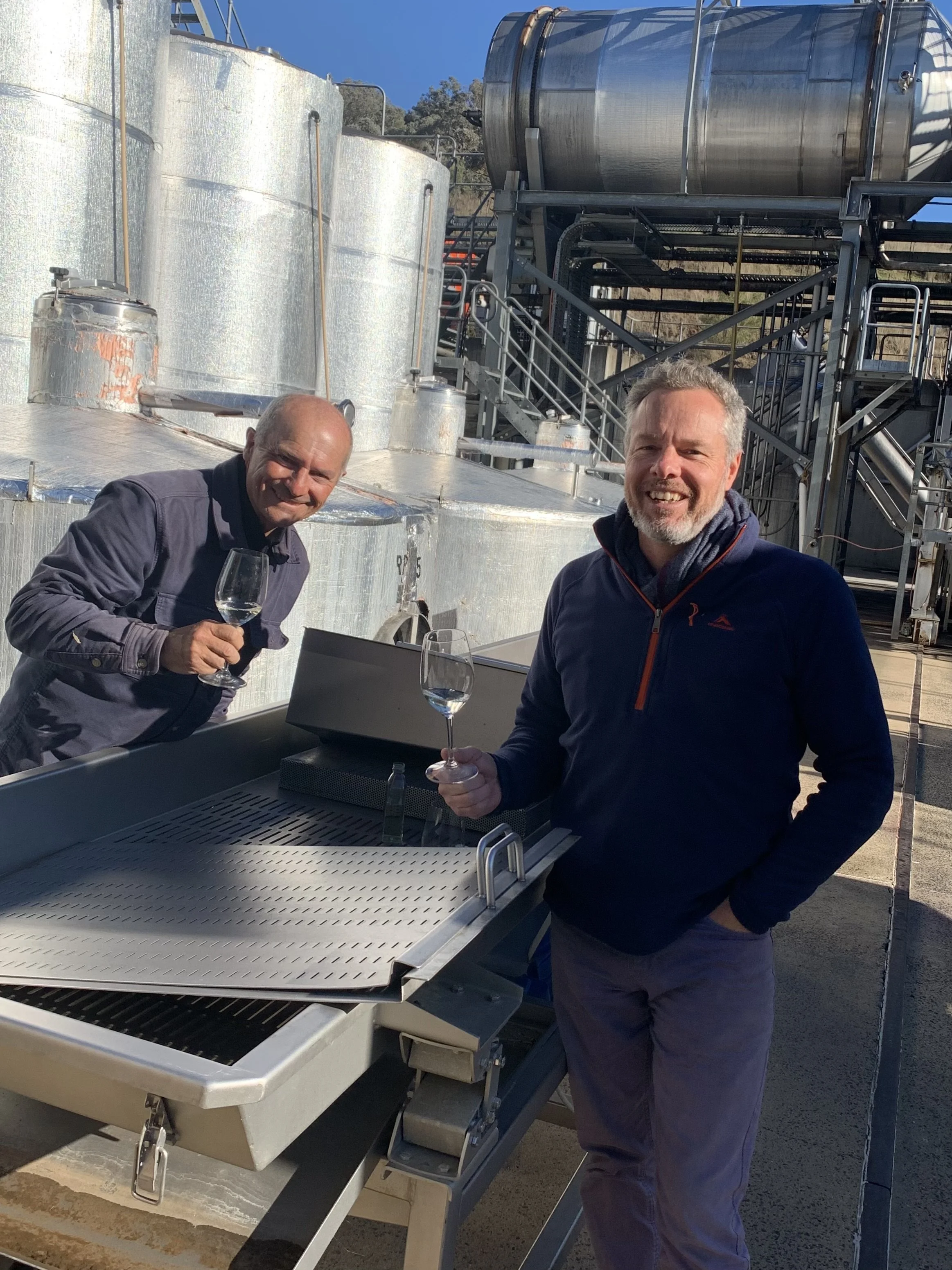 Two men enjoying glasses of wine outdoors at an industrial winery with large stainless steel tanks in the background.