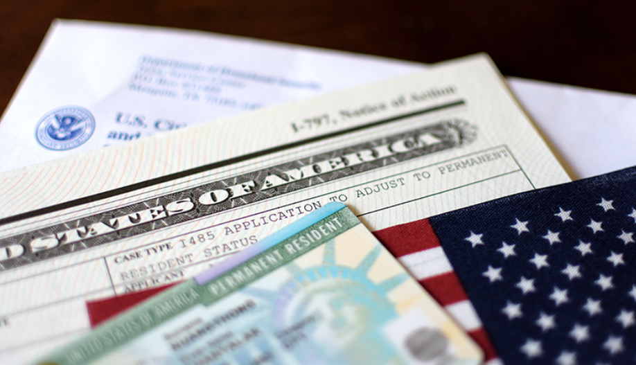 U.S. government documents, including a U.S. passport, a green card, a social security card, and an American flag, arranged on a wooden surface.