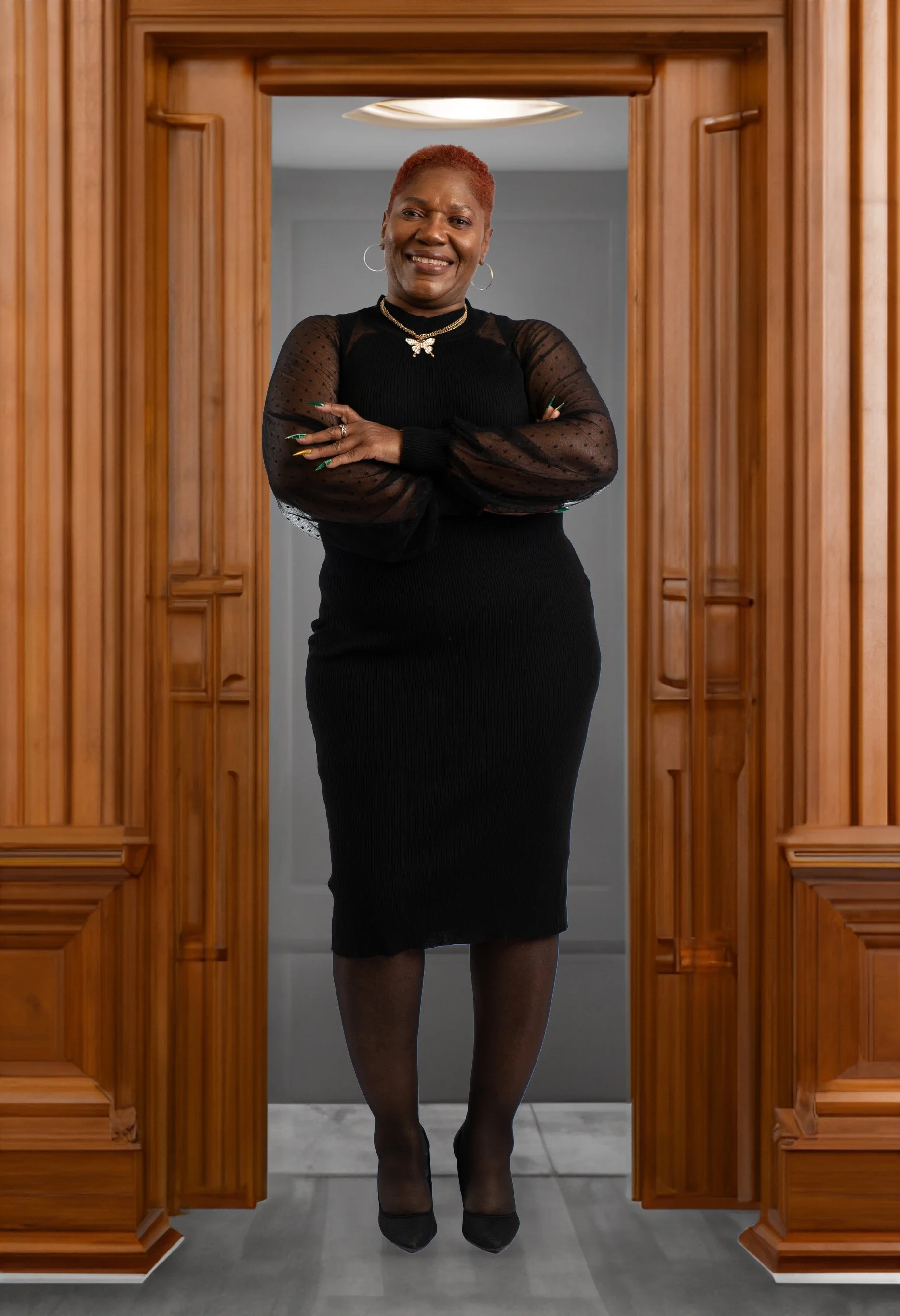 A woman standing with crossed arms, smiling, wearing a black dress with sheer polka dot long sleeves, black heels, jewelry, and earrings, framed by wooden doorways.