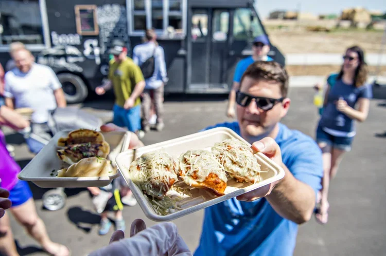 Person handing pizza and sandwiches to the camera during an outdoor gathering with a food truck and several people in the background.